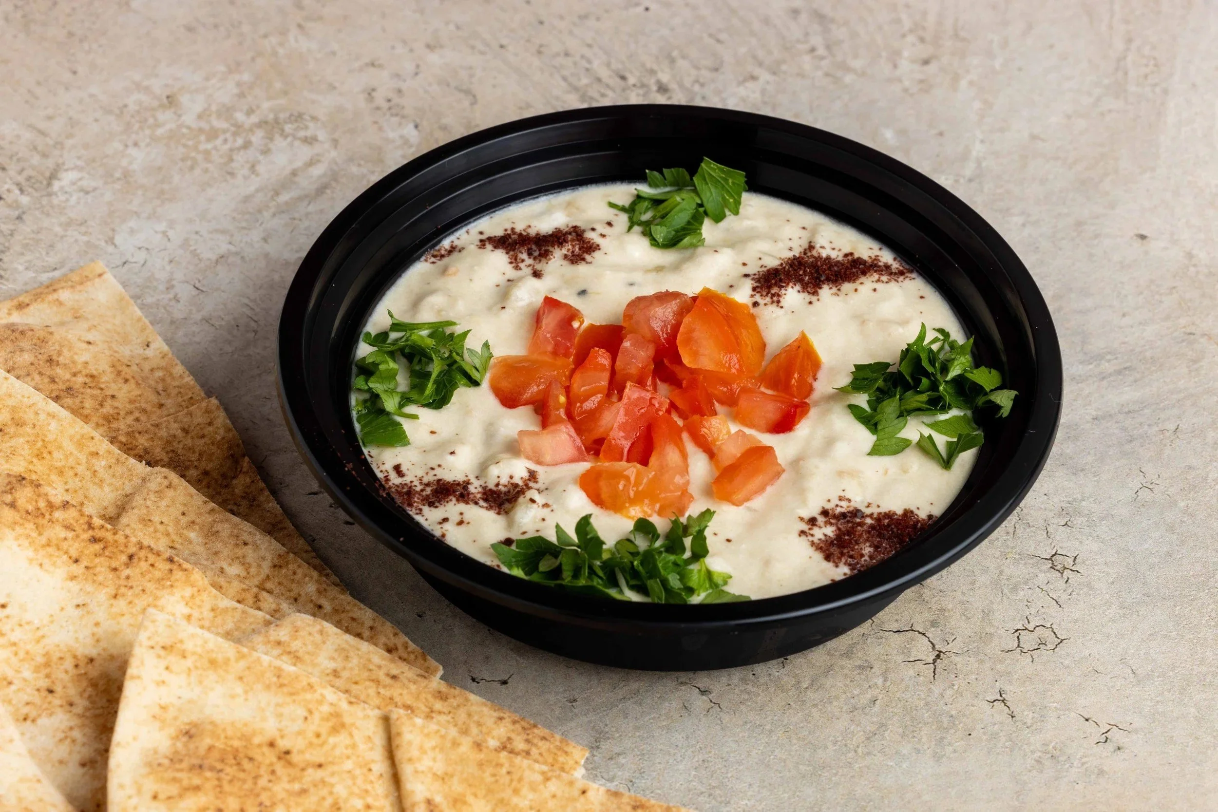 A bowl of creamy dip garnished with chopped tomatoes, fresh green herbs, and sprinkled with spices. There are folded flatbreads or chips beside it, on a gray textured surface.