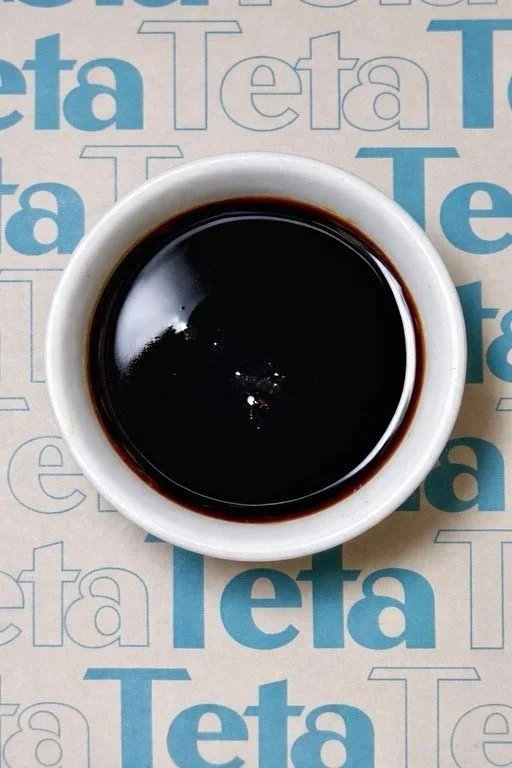 A white cup filled with black coffee on a surface with repetitive blue and brown 'Teta' text.