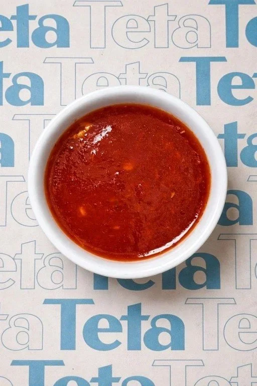 A small white bowl filled with red sauce placed on a printed paper with the word 'Teta' repeated in blue and gray letters.