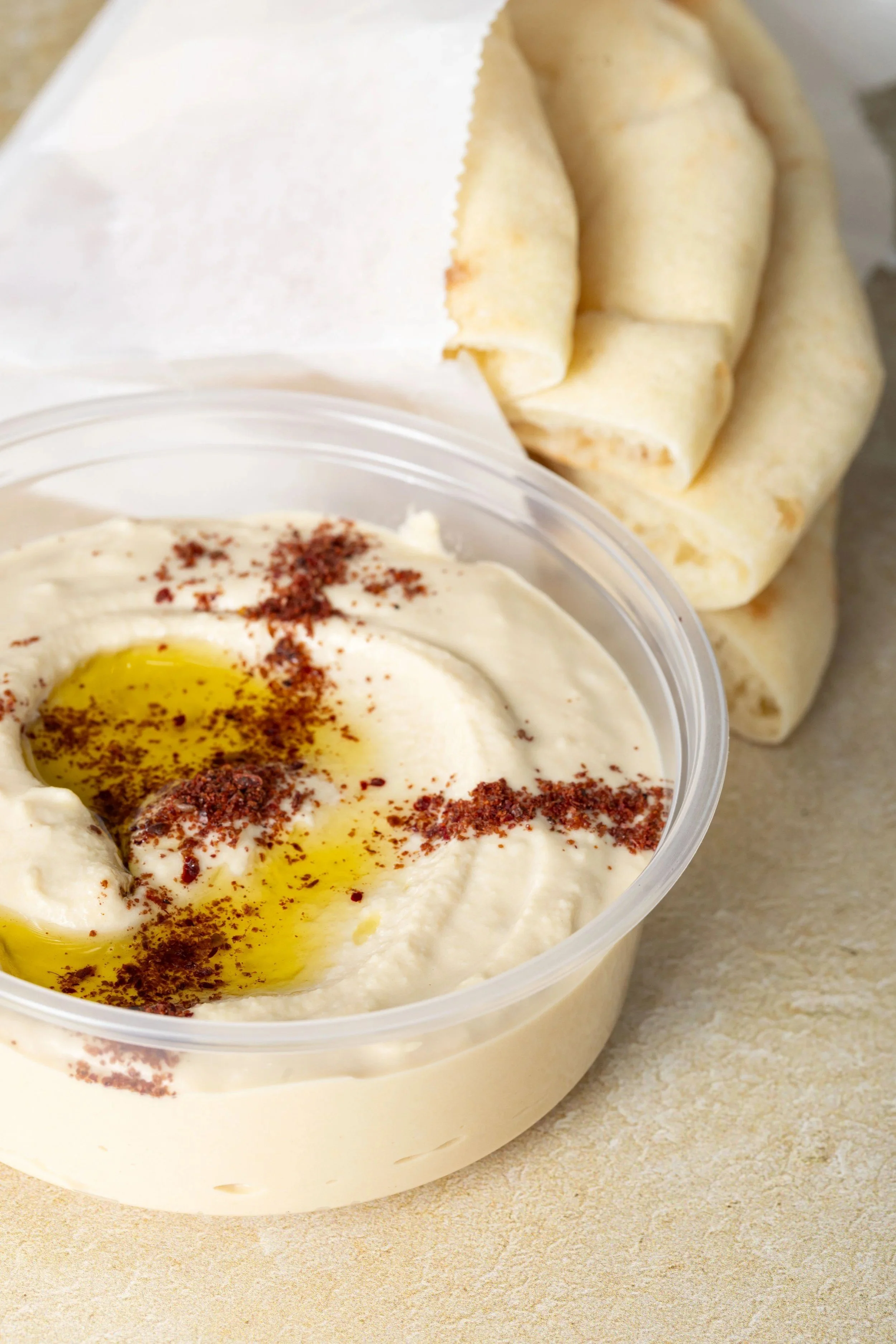 A container of creamy hummus topped with olive oil, paprika, and a sprinkle of spices, with pita bread in the background.