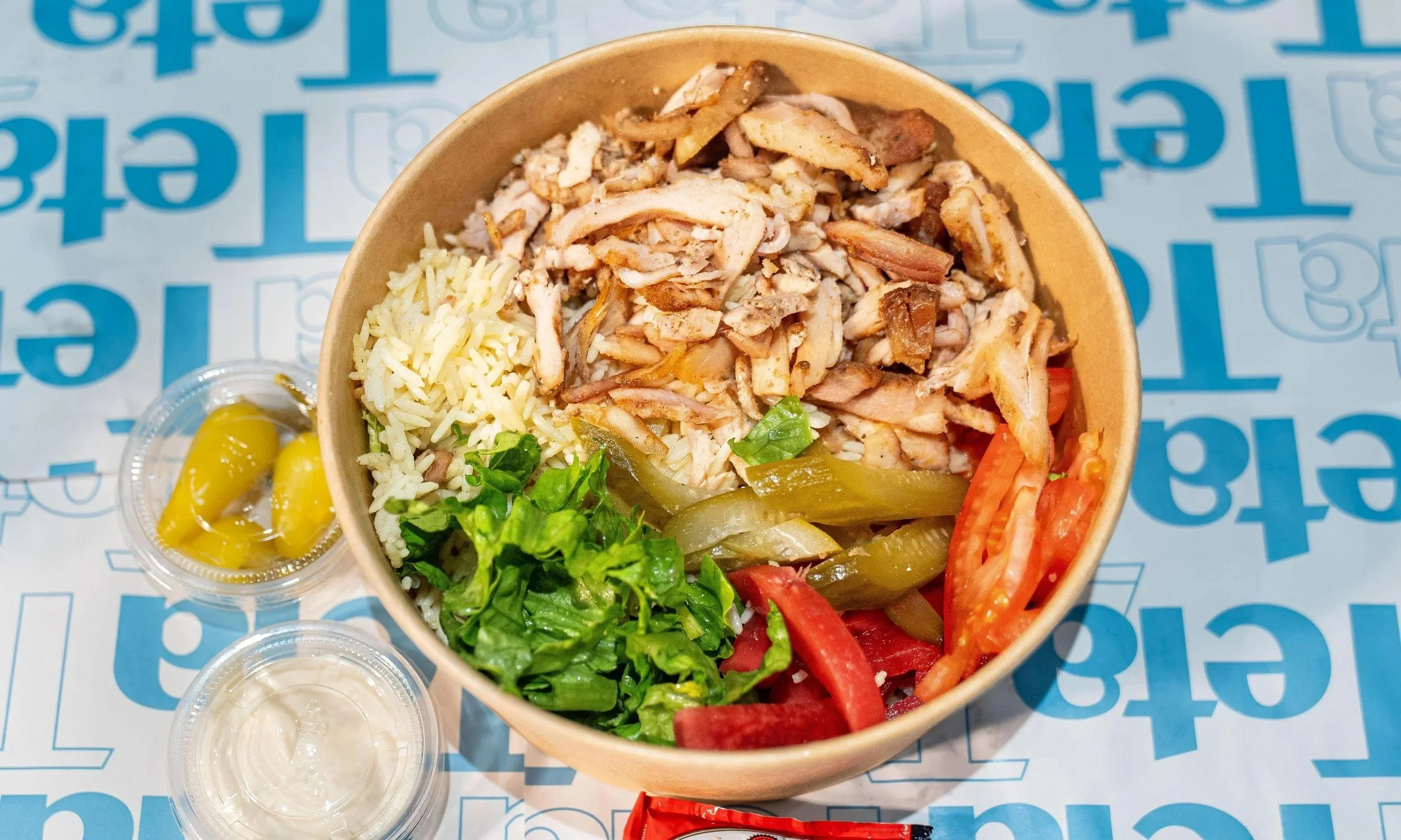 A takeout container filled with shredded chicken, rice, lettuce, tomato slices, pickled peppers, and green bell peppers, with small cups of yellow peppers and sauce on the side.
