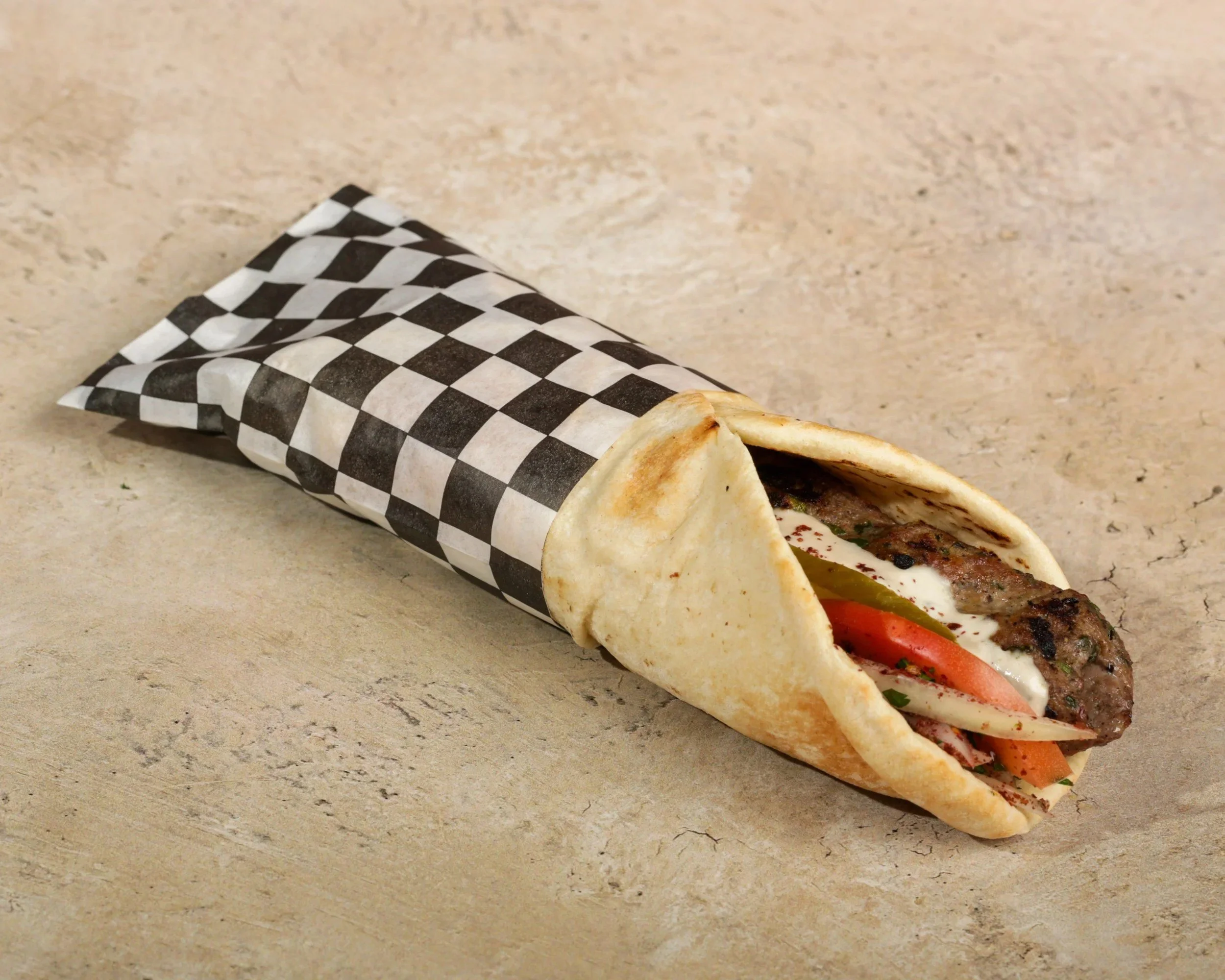 A partially unwrapped shawarma or kebab wrap with grilled meat, vegetables, and sauce in a bread bun on a beige surface.