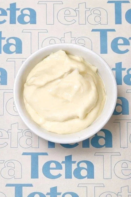 A small white bowl filled with creamy mayonnaise on a paper wrapping with a blue and white 'Teta' logo.