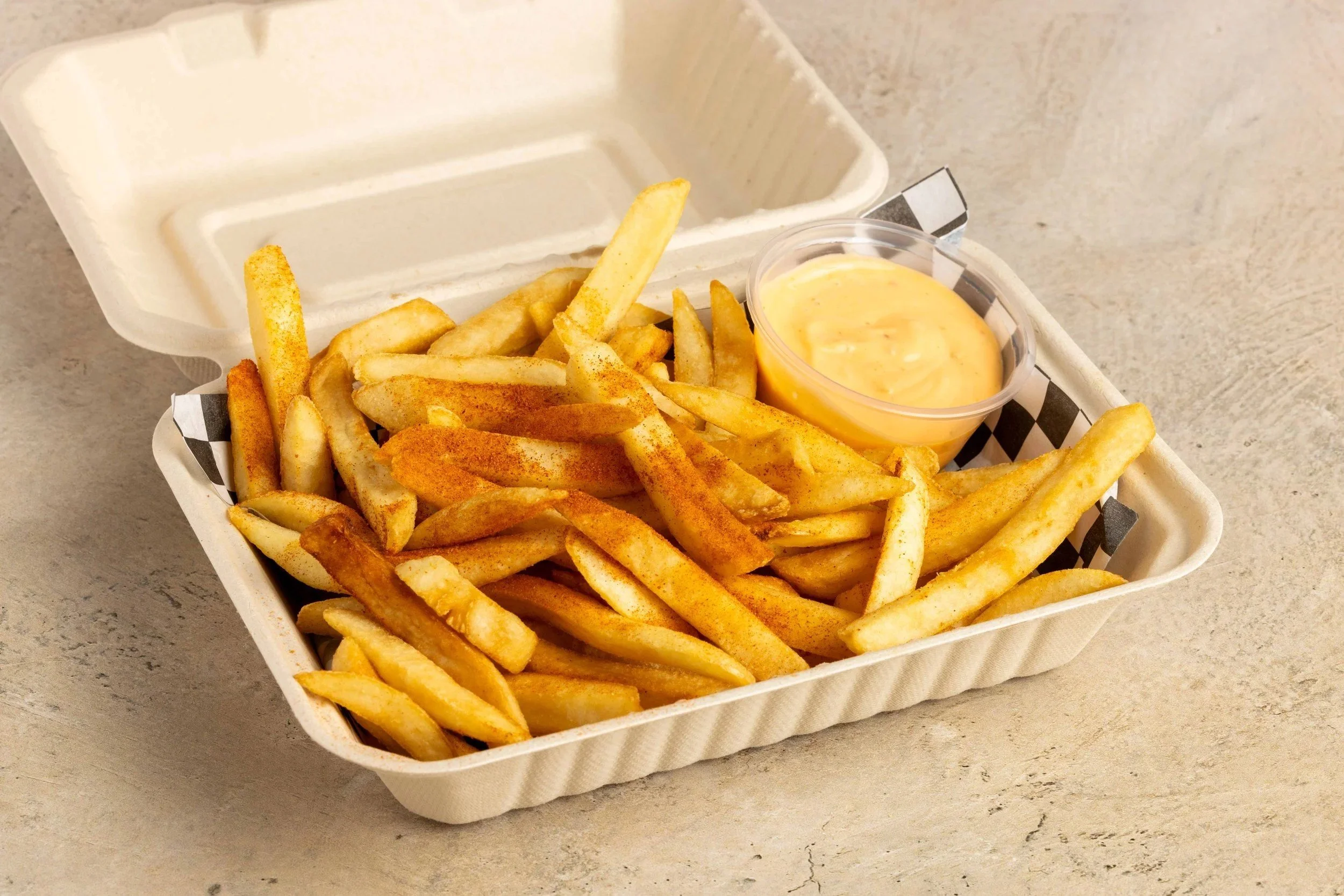 French fries seasoned with seasoning in a white takeout container, accompanied by a small cup of cheese sauce, on a beige countertop.