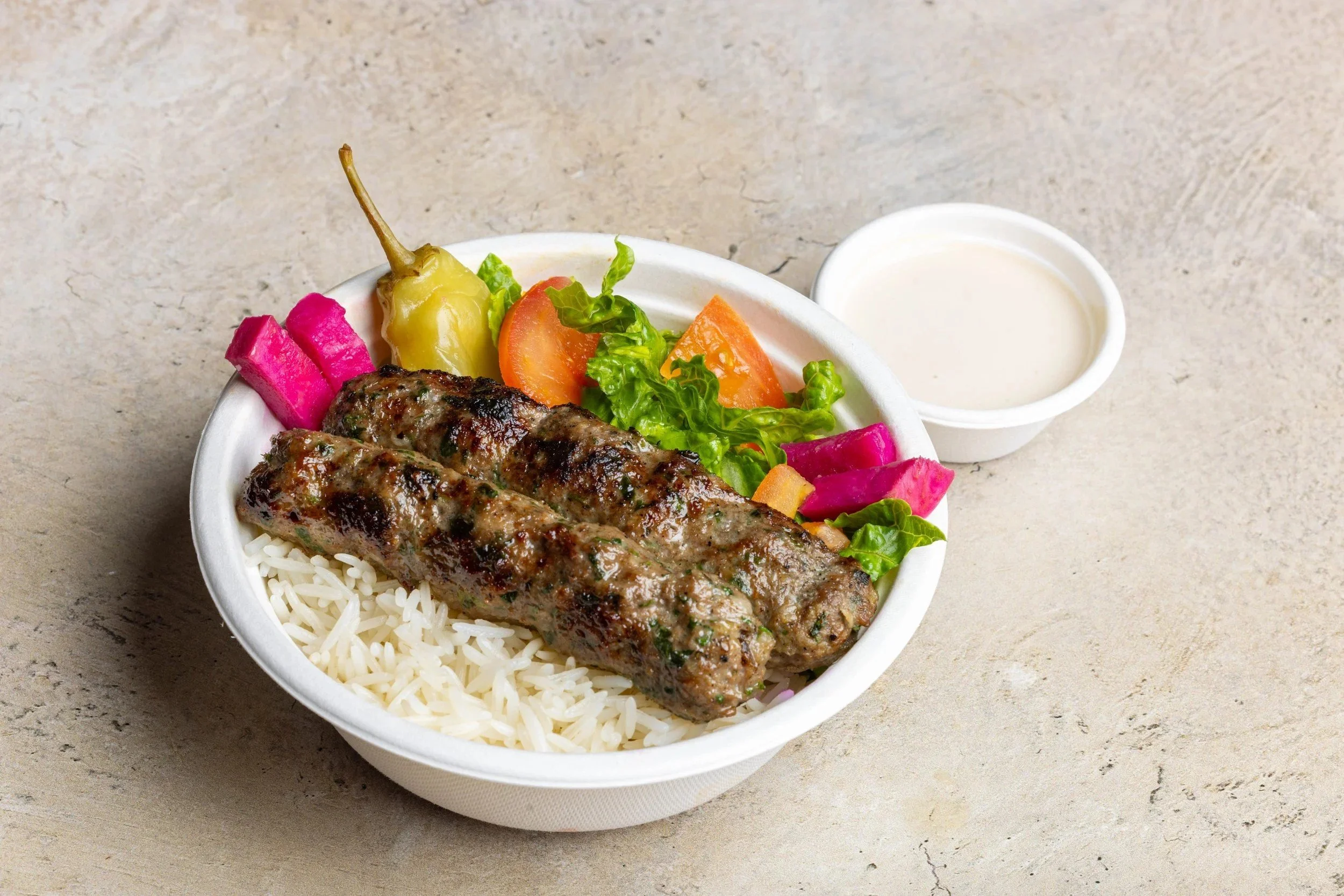 A bowl of rice with grilled kebab, fresh vegetables, pickled radish, and a side of white sauce on a gray surface.