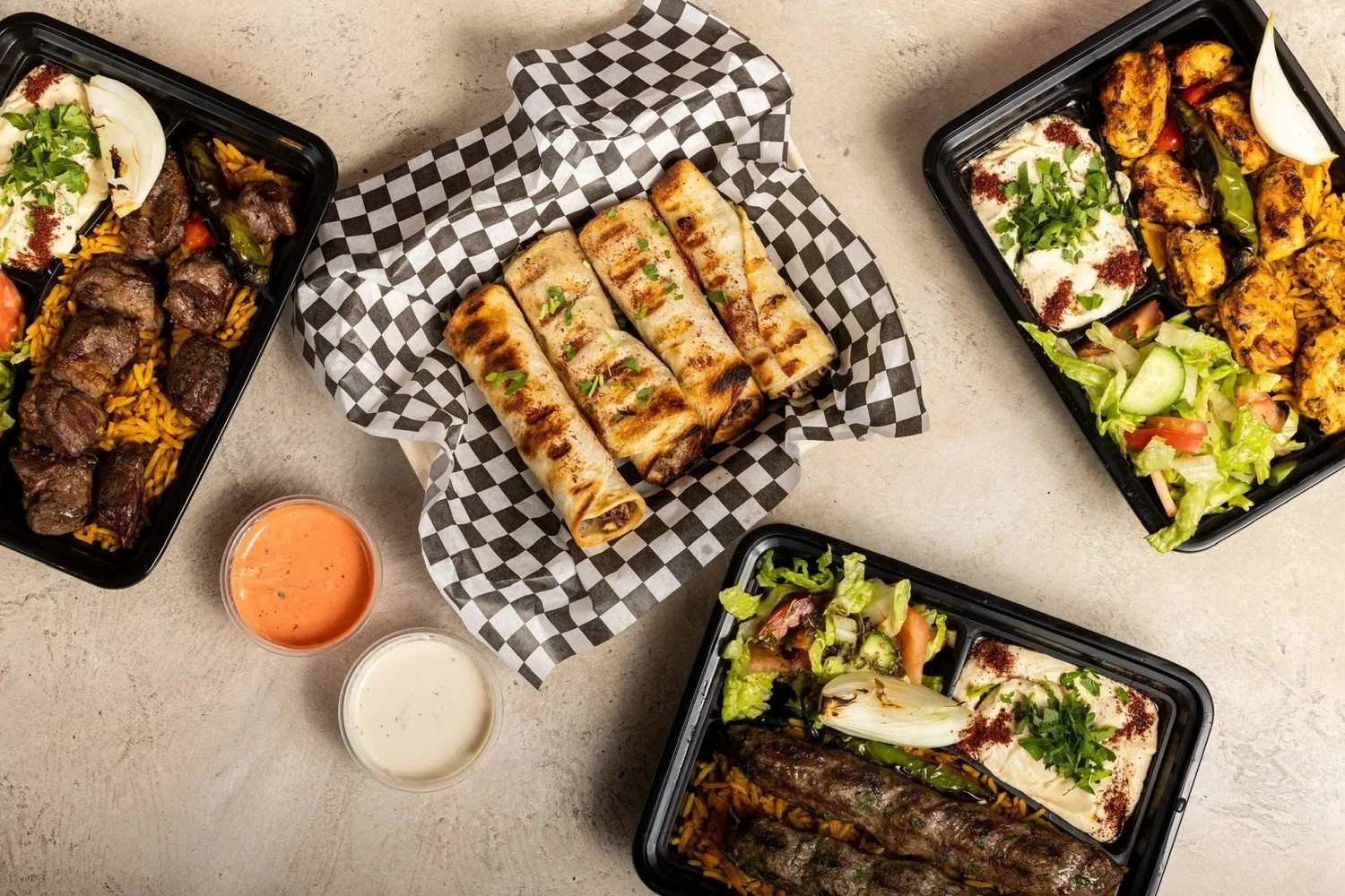 Takeout containers with grilled meats, rice, salads, and grilled vegetables, along with two dipping sauces on a neutral surface.