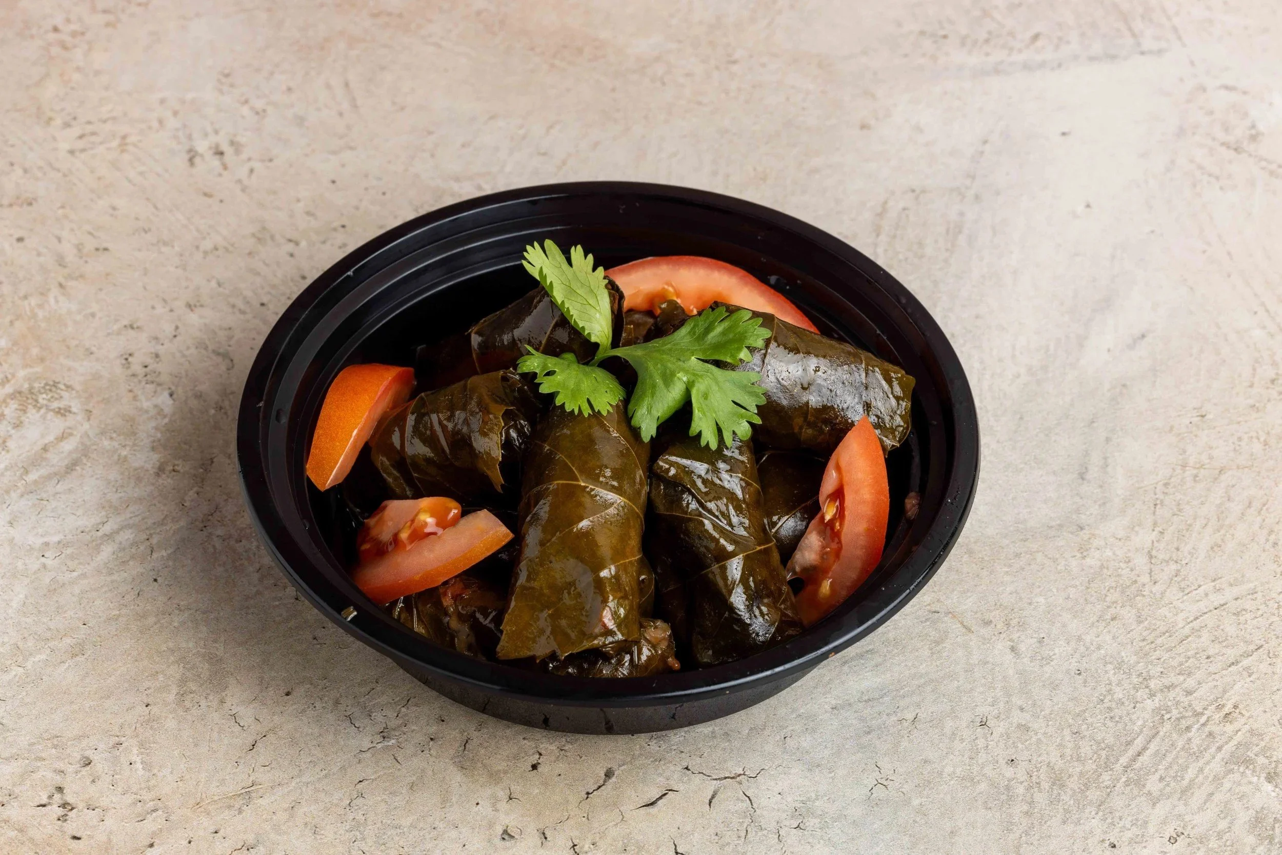 A black bowl containing stuffed grape leaves garnished with fresh cilantro and sliced tomatoes on a light-colored surface.