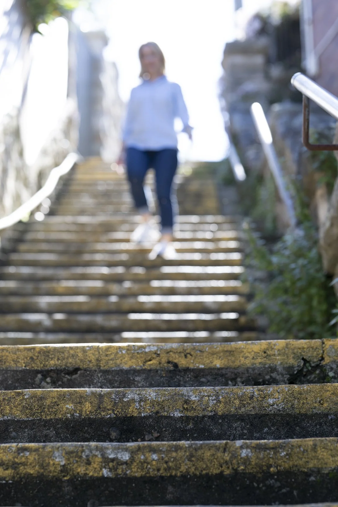 Blurred photo of a woman walking down outdoor stone stairs with metal handrails on both sides, sunlight in the background.