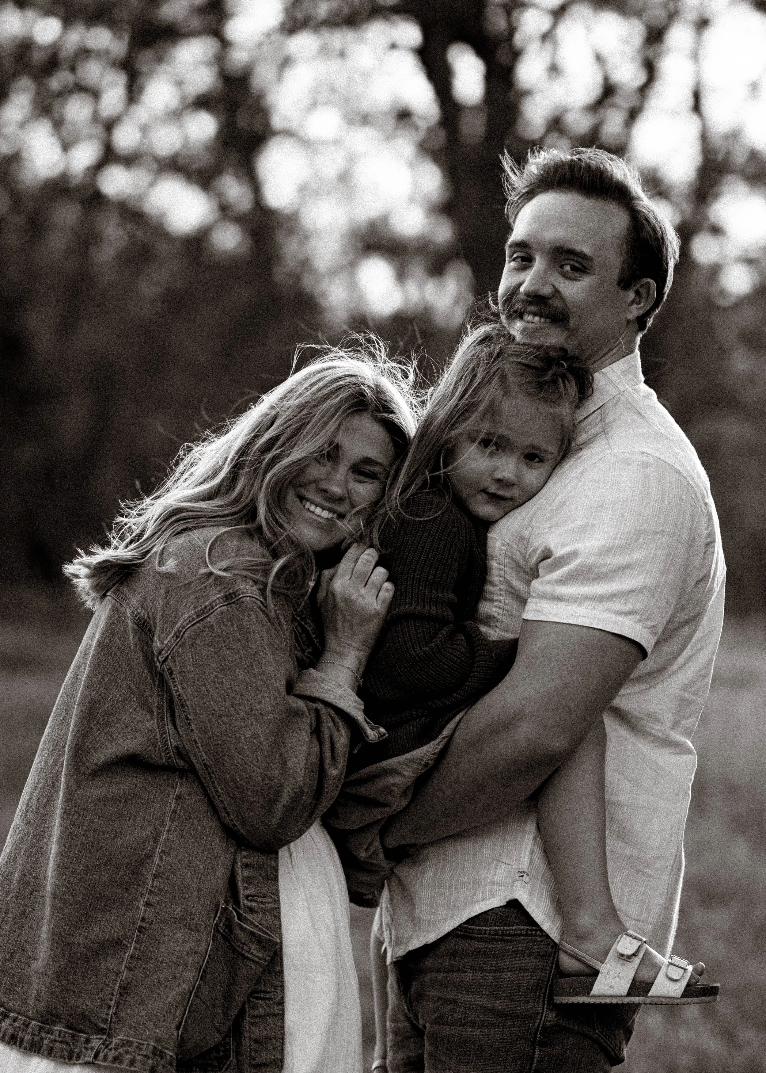 A happy family outdoors, with a man holding a young girl and a woman smiling. The family is close together, embracing and smiling at the camera, with trees in the background.