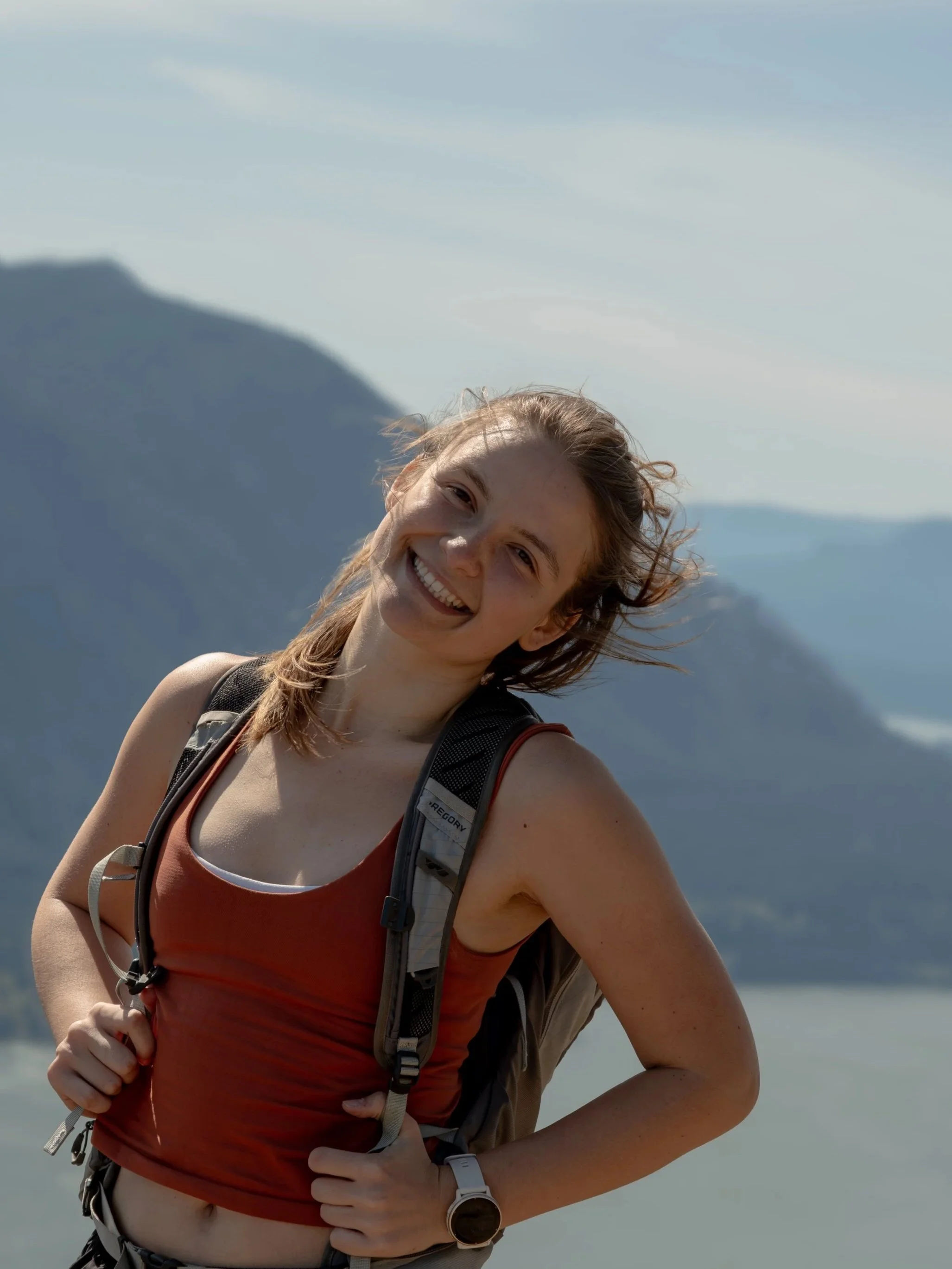 A young woman smiling while hiking outdoors on a sunny day, wearing a red tank top, carrying a backpack, with mountains and sky in the background.