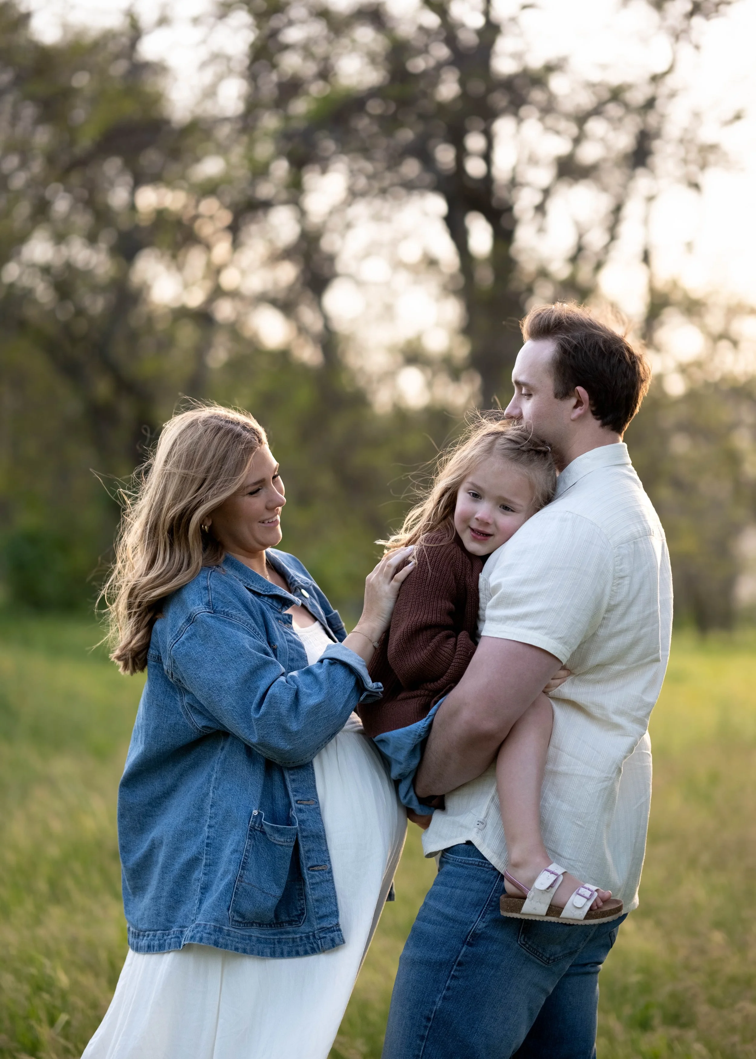 A family of four standing outdoors in a grassy field with trees in the background, sharing a moment of affection and joy.