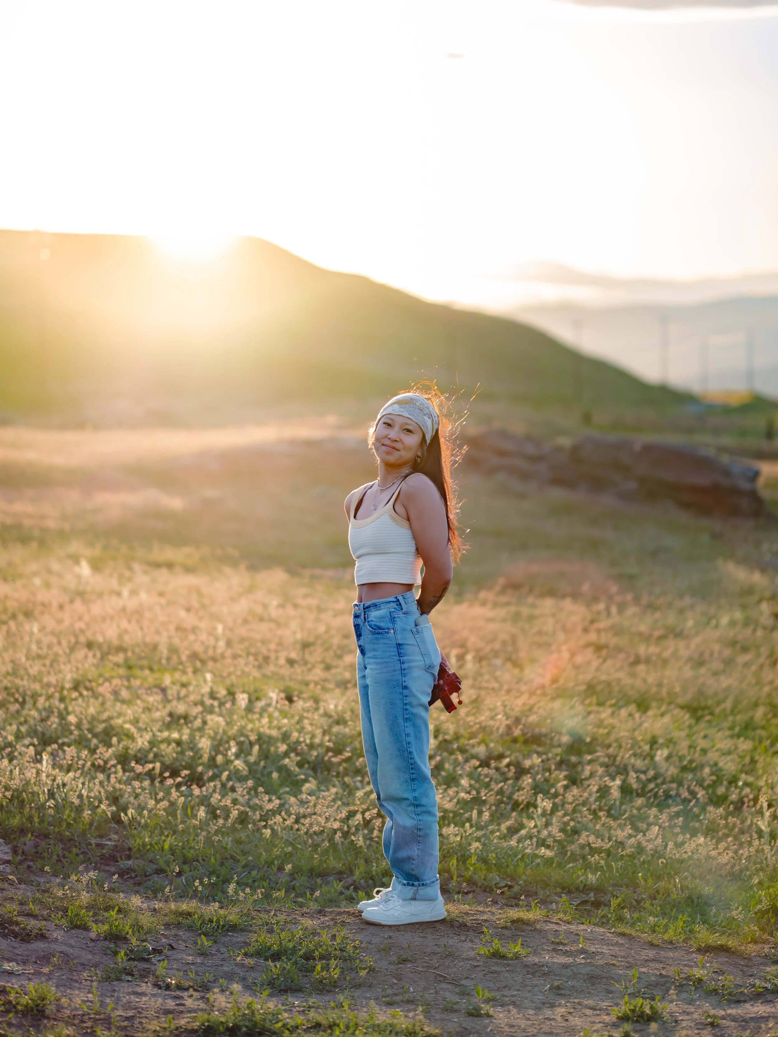 A young woman stands outdoors in a field at sunset, wearing a white crop top, blue jeans, white sneakers, and a headband, with a hill in the background.