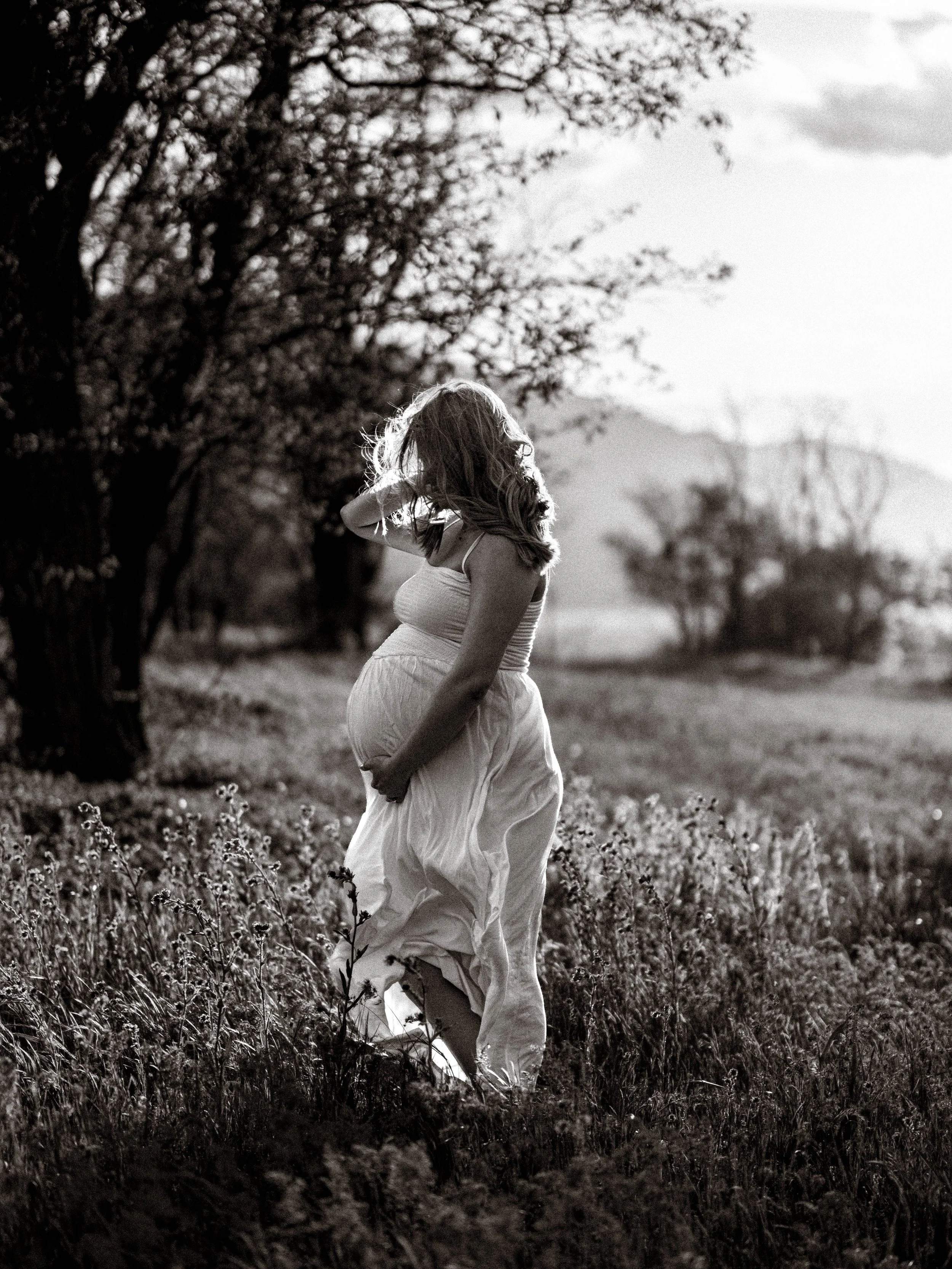 A black and white photo of a woman with long hair standing in a field of tall grass and flowers, touching her hair, with trees and mountains in the background.
