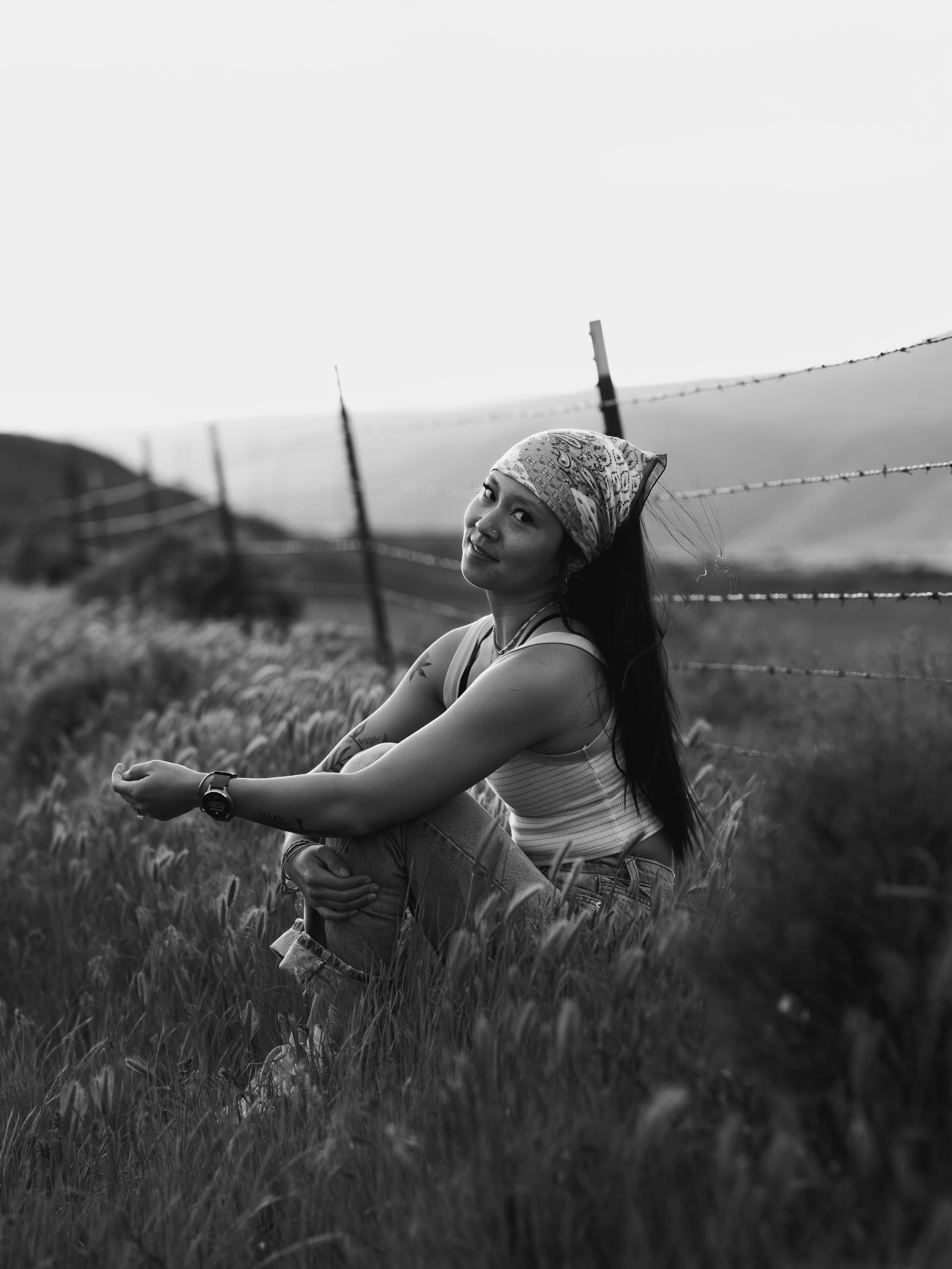 A woman with a bandana on her head sitting in a field of tall grass, smiling, with a fence and hills in the background.