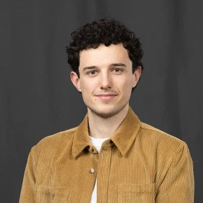 A young man with curly dark hair, wearing a tan jacket over a white shirt, standing in front of a plain dark gray background.