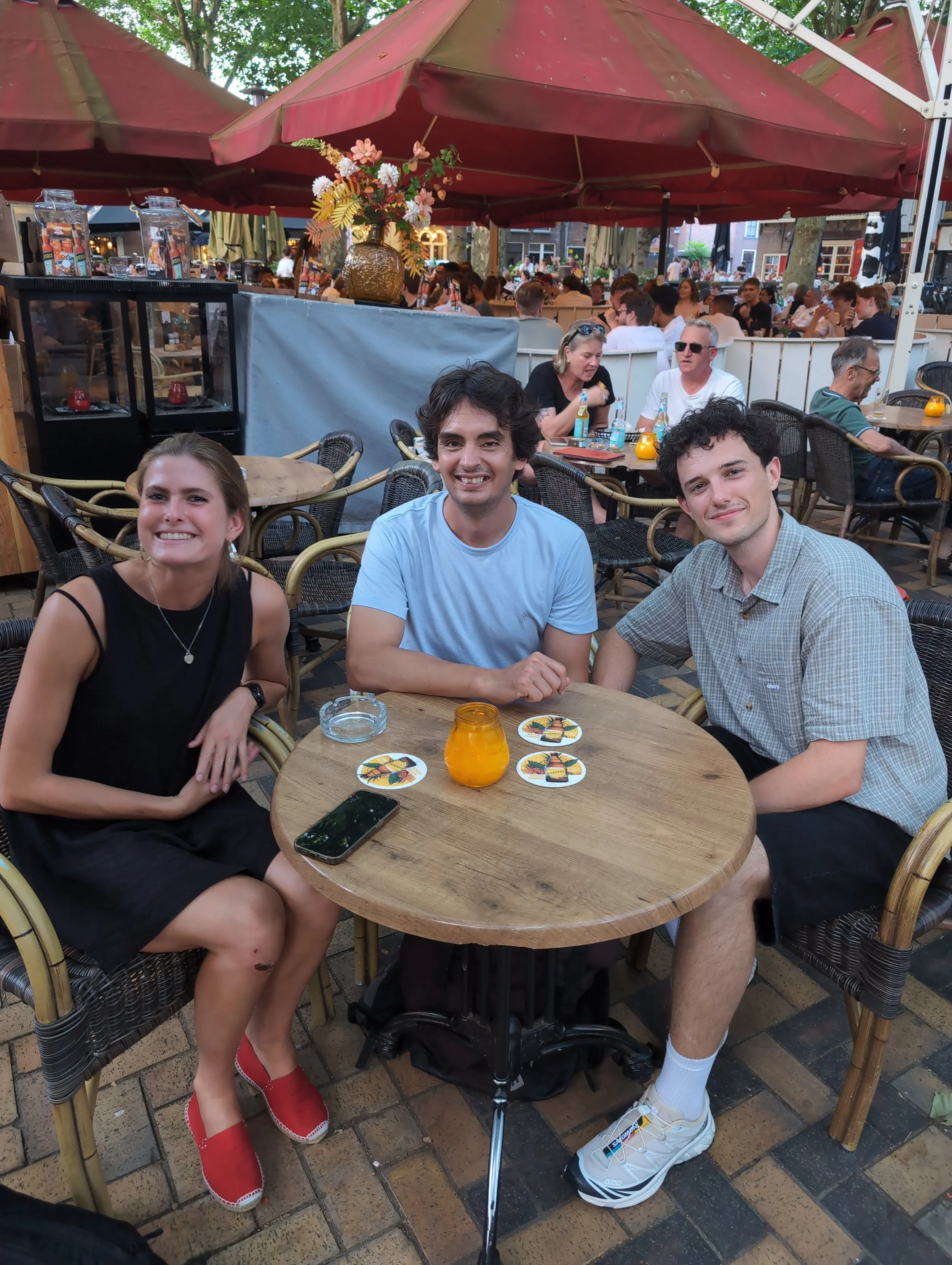 Three young adults smiling at a round outdoor table with colorful coasters and a glass of orange drink, seated under a large red patio umbrella, with many people in the background at a busy outdoor restaurant or cafe.