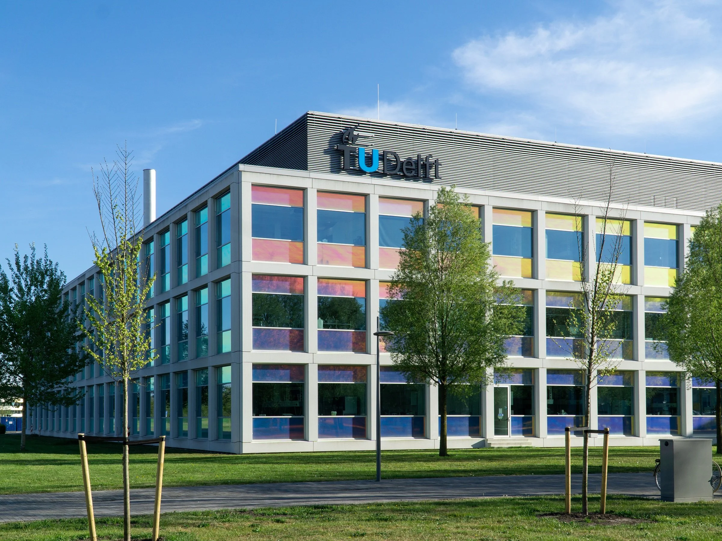 Modern office building with colorful windows, signage reading 'TUDelft', surrounded by trees and a green lawn under a partly cloudy sky.