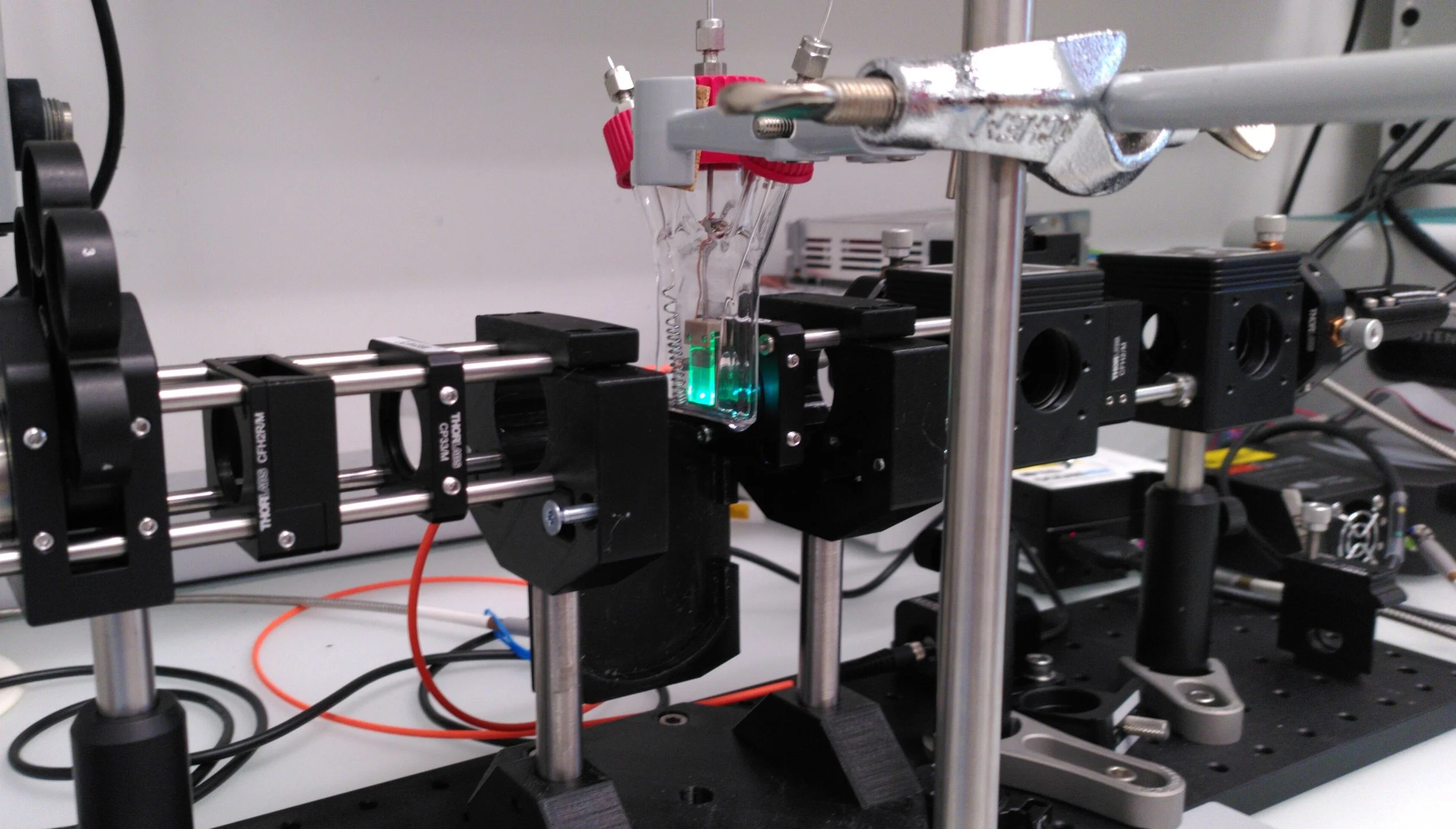 Laboratory setup with optical and mechanical components, including a glass container with liquid and green light, connected to metal rods and mounts for scientific experiments.