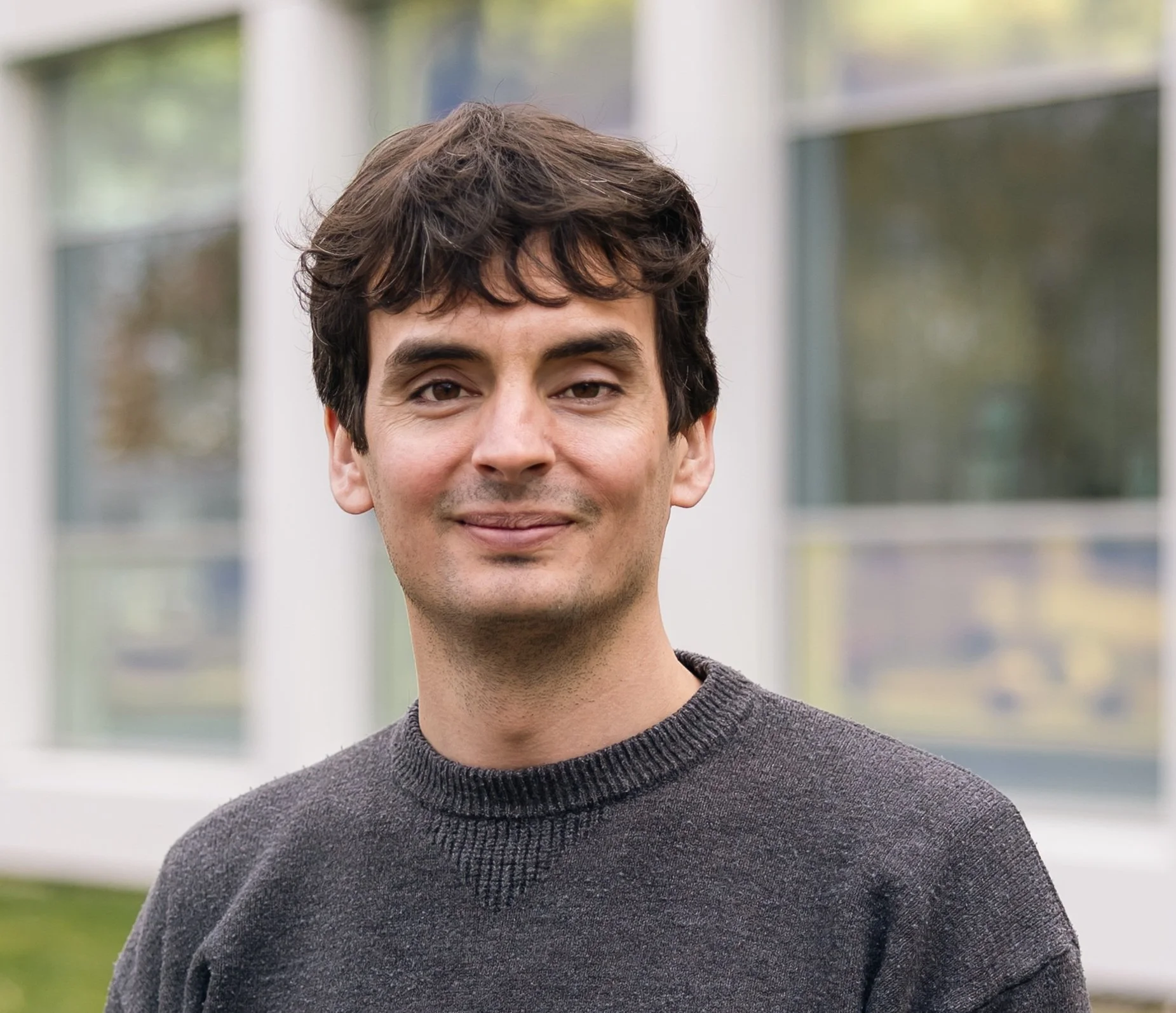 Close-up of a young man with dark wavy hair, smiling softly, wearing a gray sweater, outdoors with blurred background of windows and greenery.