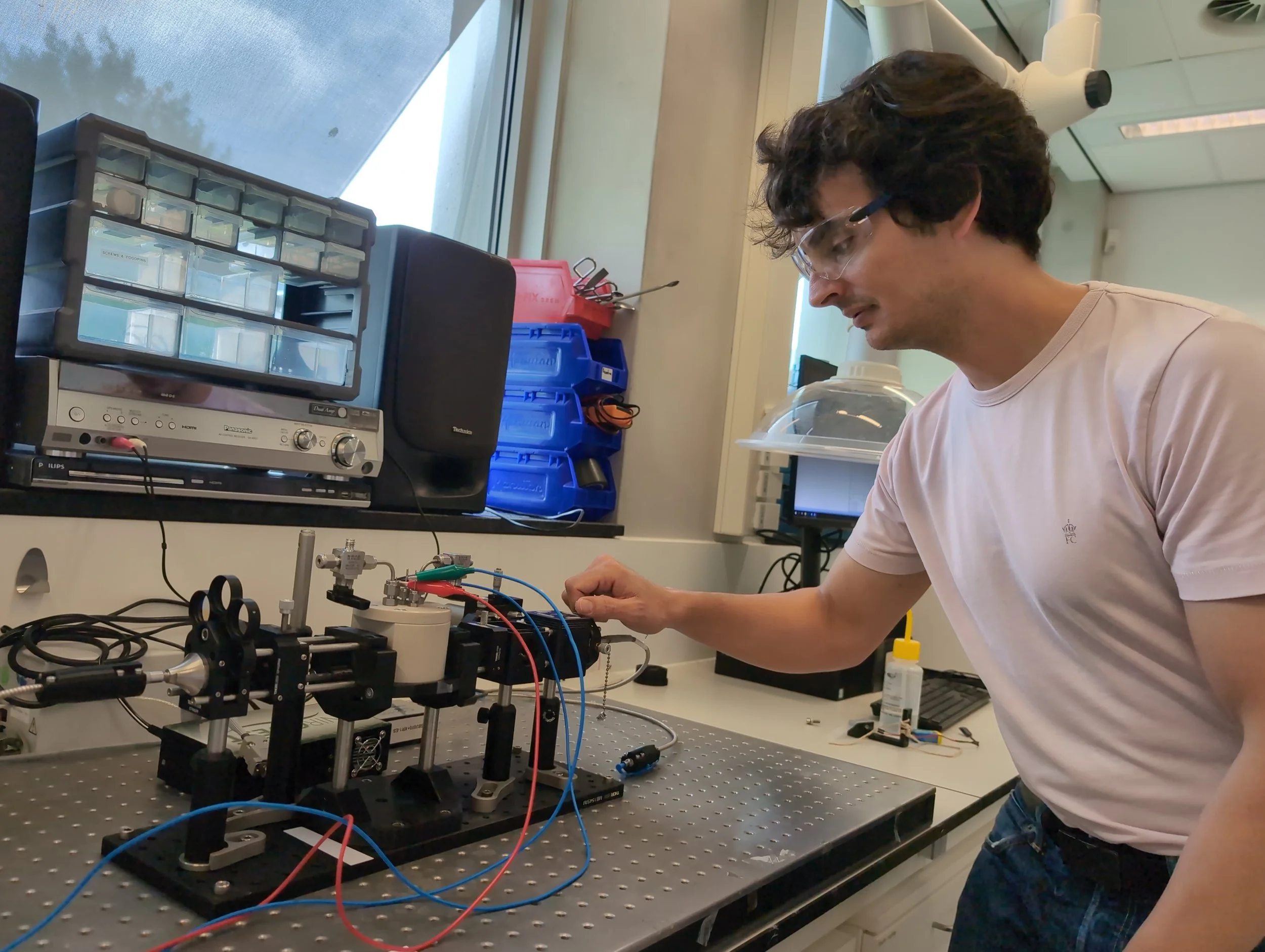 A man with glasses working on a scientific experiment at a lab bench. The desk has scientific equipment, wires, and components, with a window and storage boxes in the background.