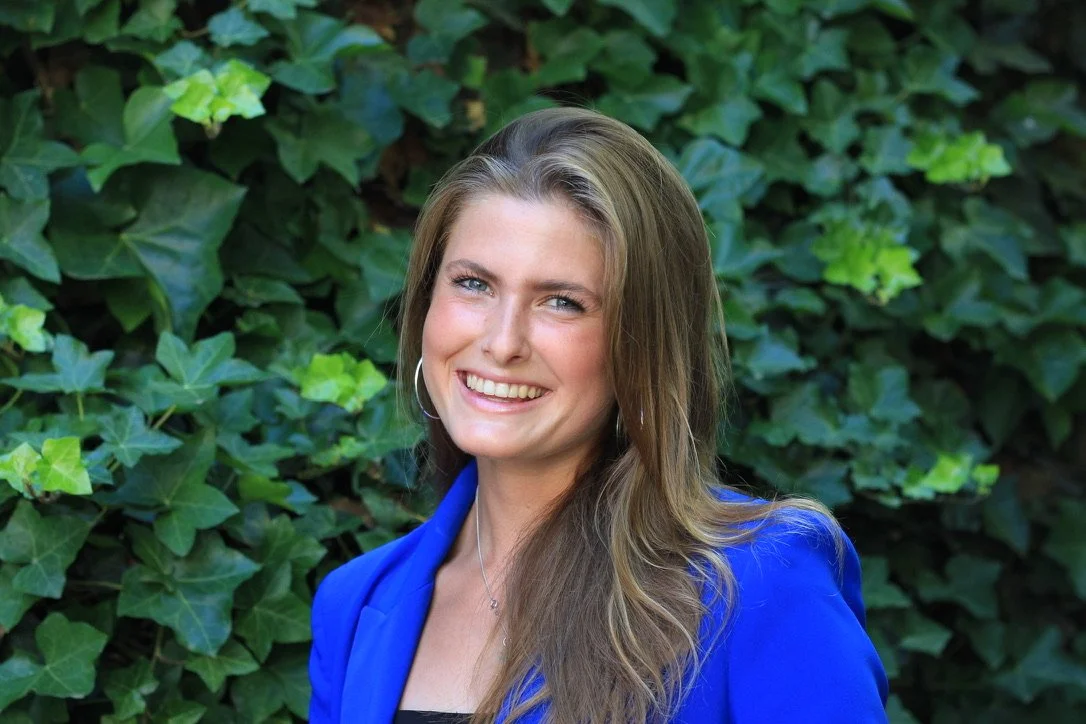 A woman smiling outdoors, wearing a blue blazer and silver jewelry, with green leafy plants in the background.