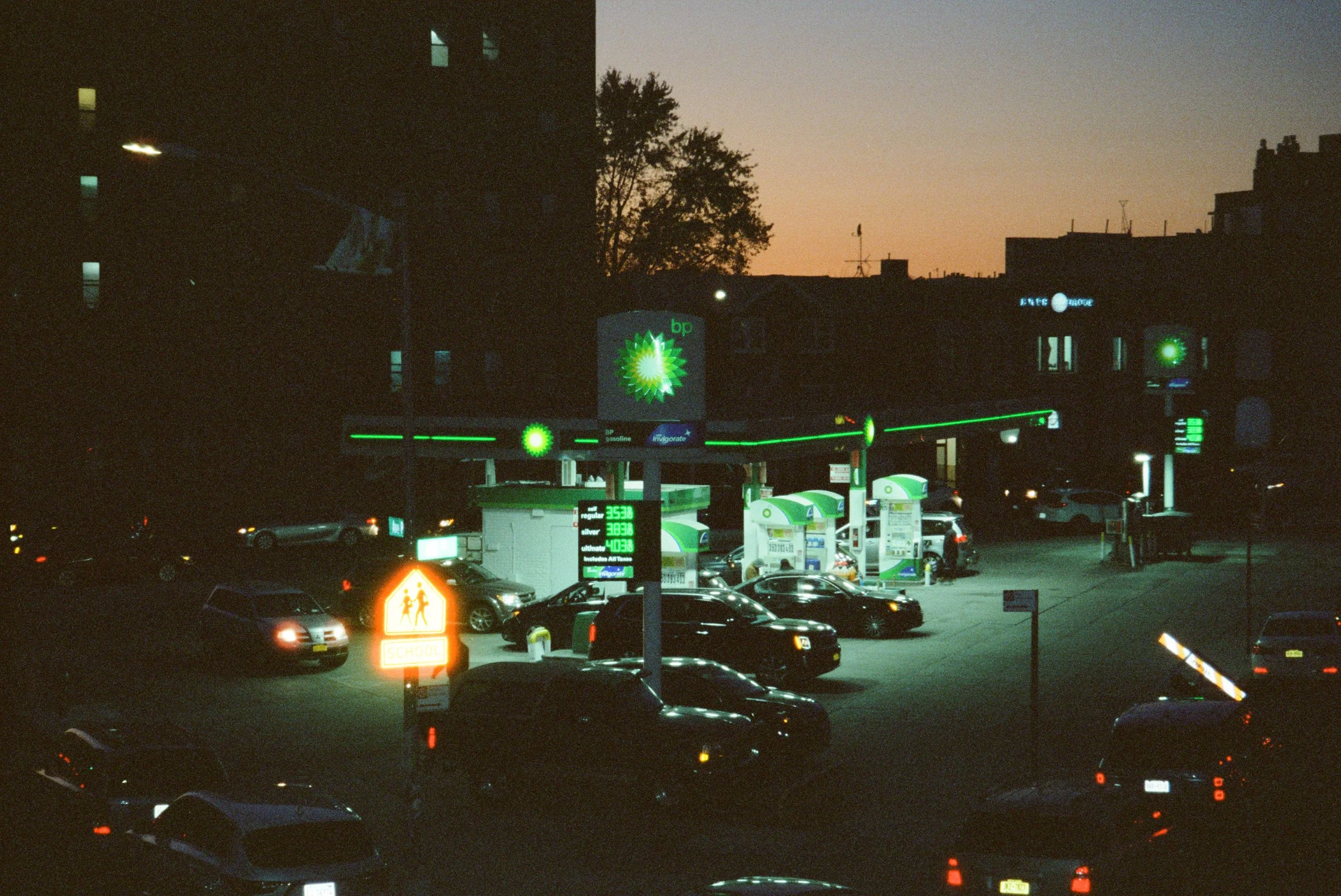 A gas station with illuminated green signage and pumps, filled with cars at dusk, with a dark building and silhouette of trees in the background, and a sunset sky.