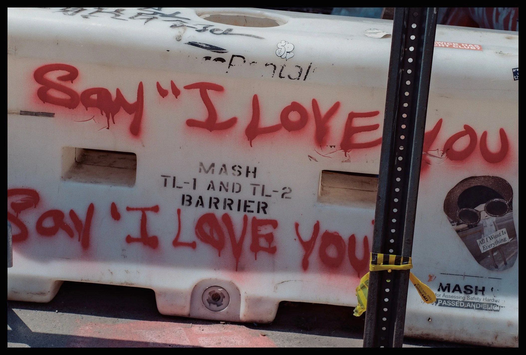 A white barricade with red spray-painted graffiti that reads 'SAY IT LOVE YOU,' includes a small drawing of a cloud with a smiling face and hearts, and is secured with black and yellow tape. A black vertical pole with white dots is on the right side,