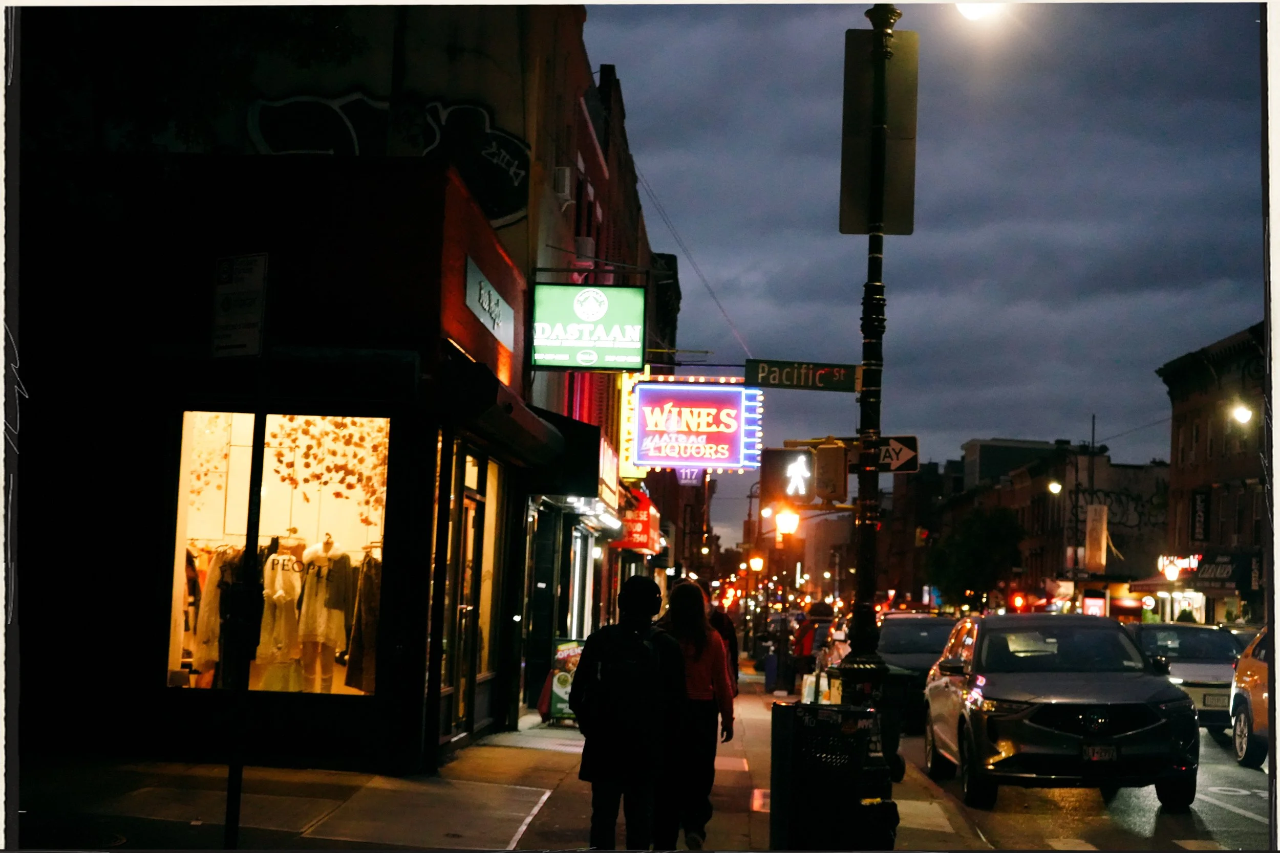 Nighttime street scene with illuminated signs, pedestrians, cars, storefronts, and a cloudy sky.
