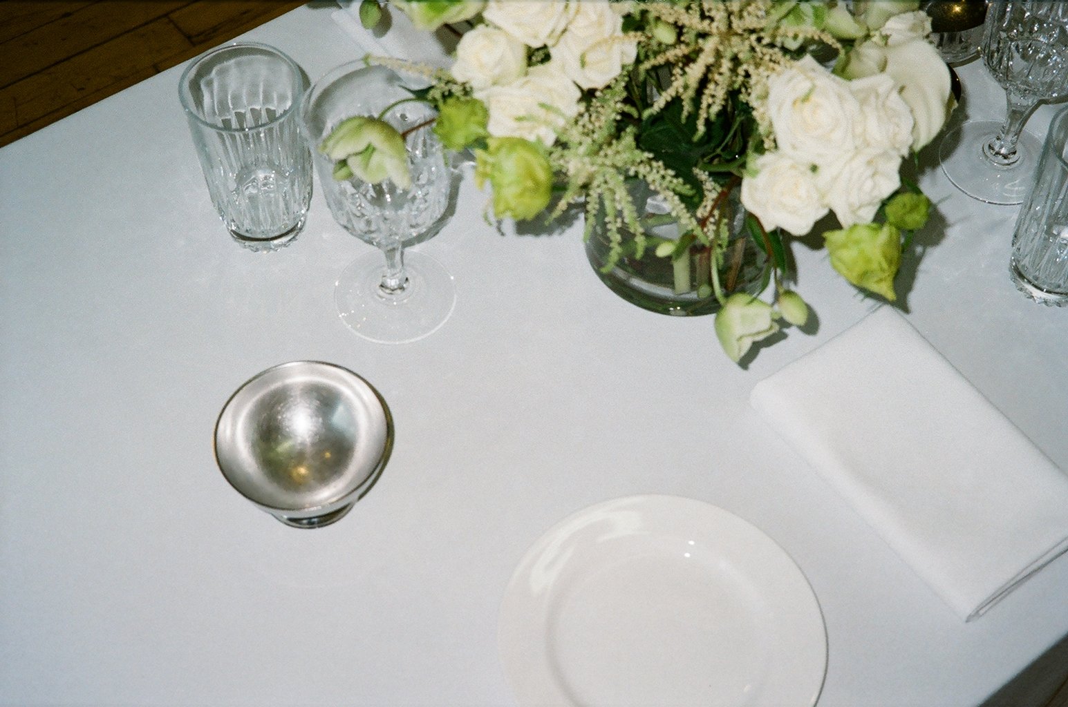 Elegant table setting with a white tablecloth, a flower arrangement in a glass vase, empty wine glasses, a metallic bowl, and a white napkin.