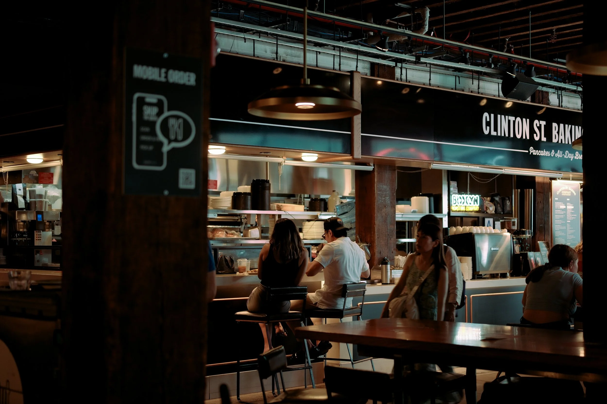 Interior of a busy café with customers sitting at the counter and tables, visible kitchen equipment, and a sign reading 'Clinton St. Baking' in the background, with industrial lighting and exposed ceiling pipes.