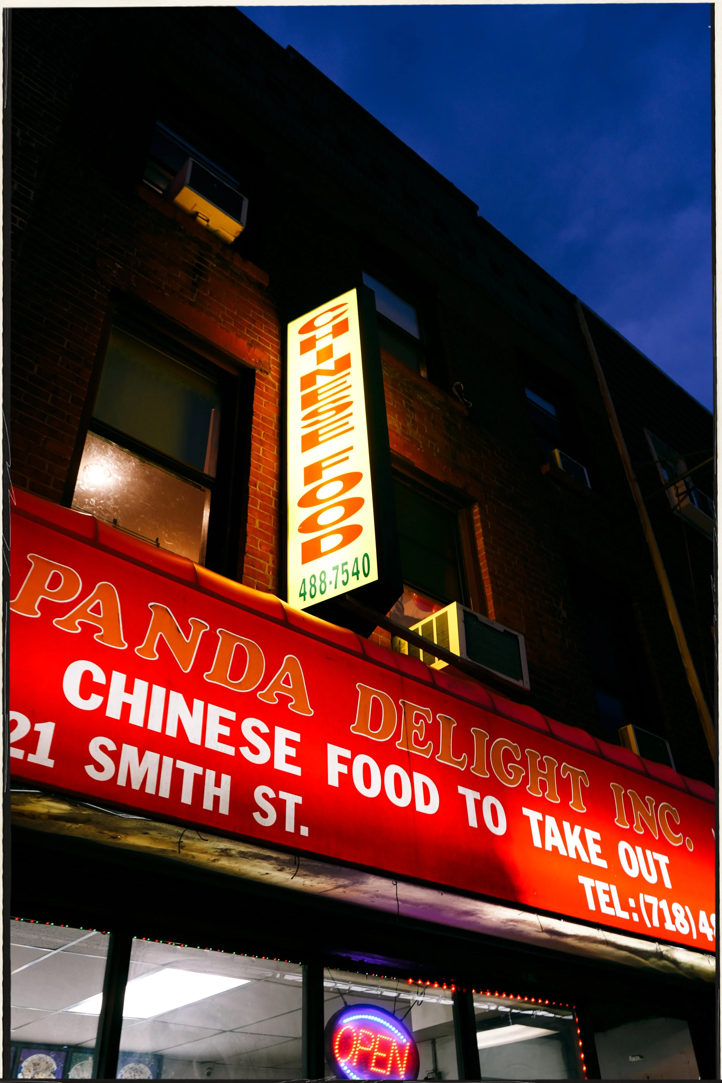 Nighttime view of a brick building with a vertical illuminated sign reading 'Chinese Food' and a red storefront awning advertising Chinese desserts and food to take out. Inside the restaurant, a neon 'Open' sign is visible.