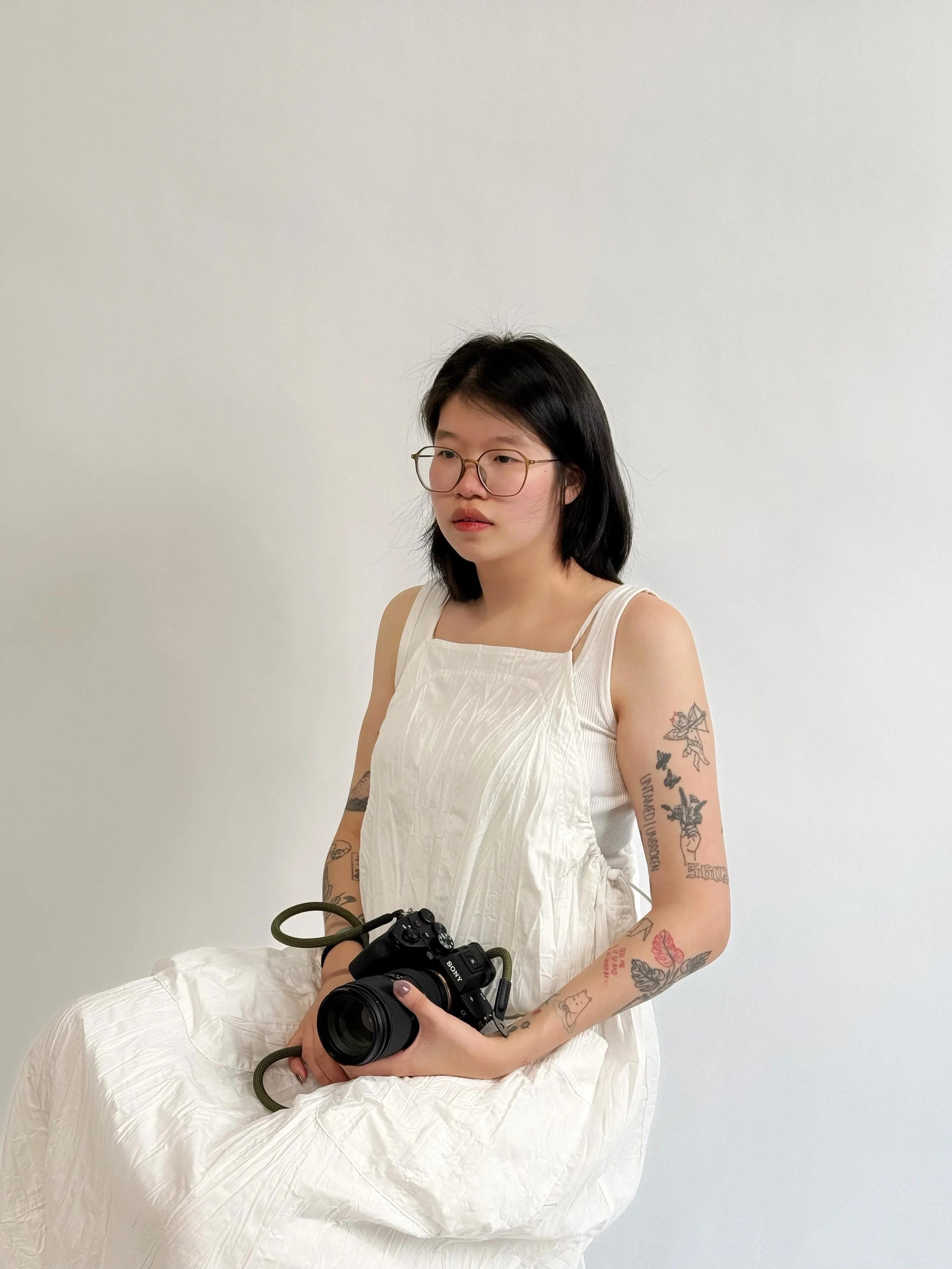 Young woman with glasses and tattoos sitting on a white chair, holding a camera, against a plain white wall background.