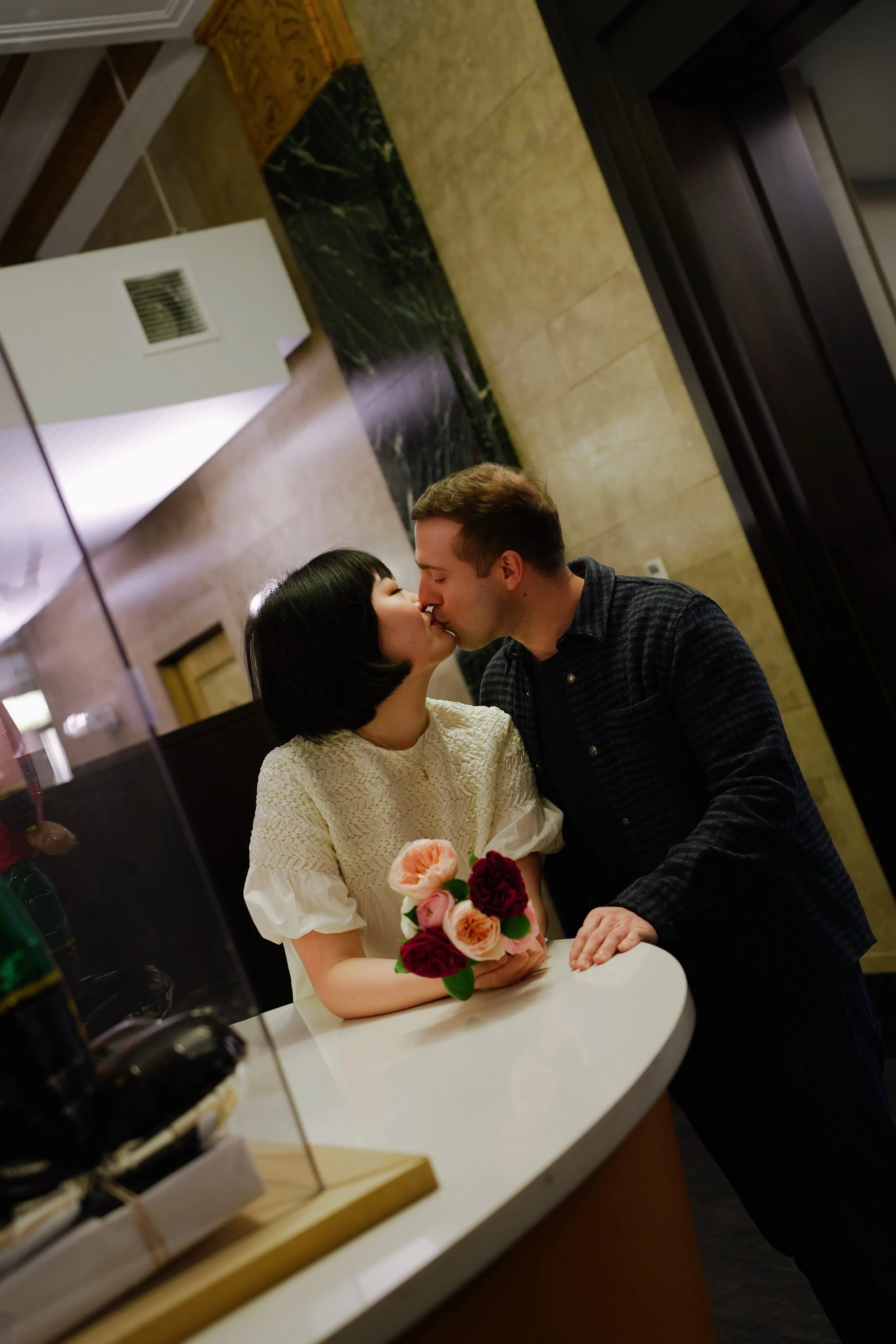 A couple kissing across a small round white table, with a bouquet of pink and dark red roses, inside a warmly-lit room with a marble and stone wall background.