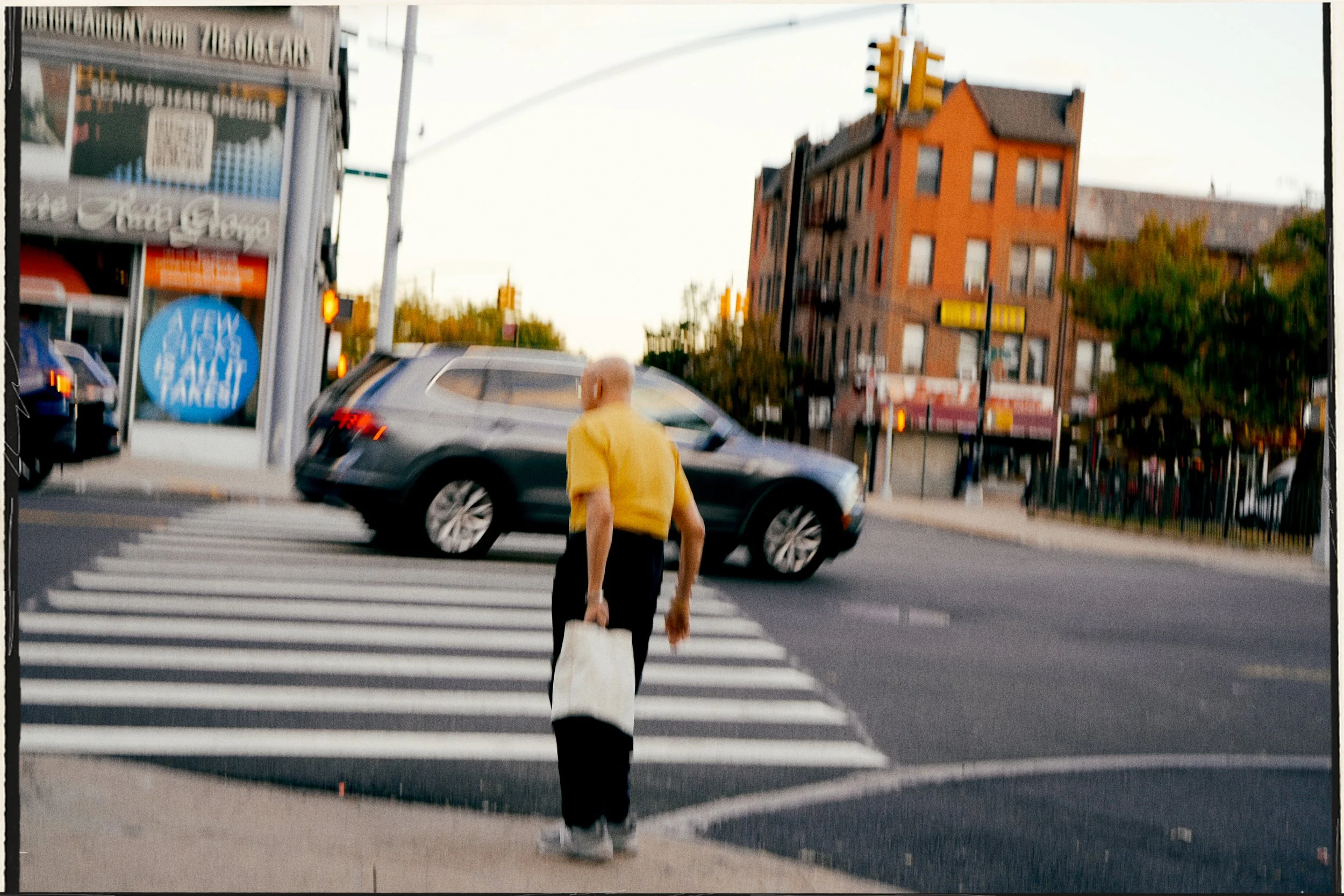 A person in a yellow shirt and dark pants crossing the street at a crosswalk with a shopping bag in hand, with a car waiting at the intersection. In the background, there are buildings and a traffic light.