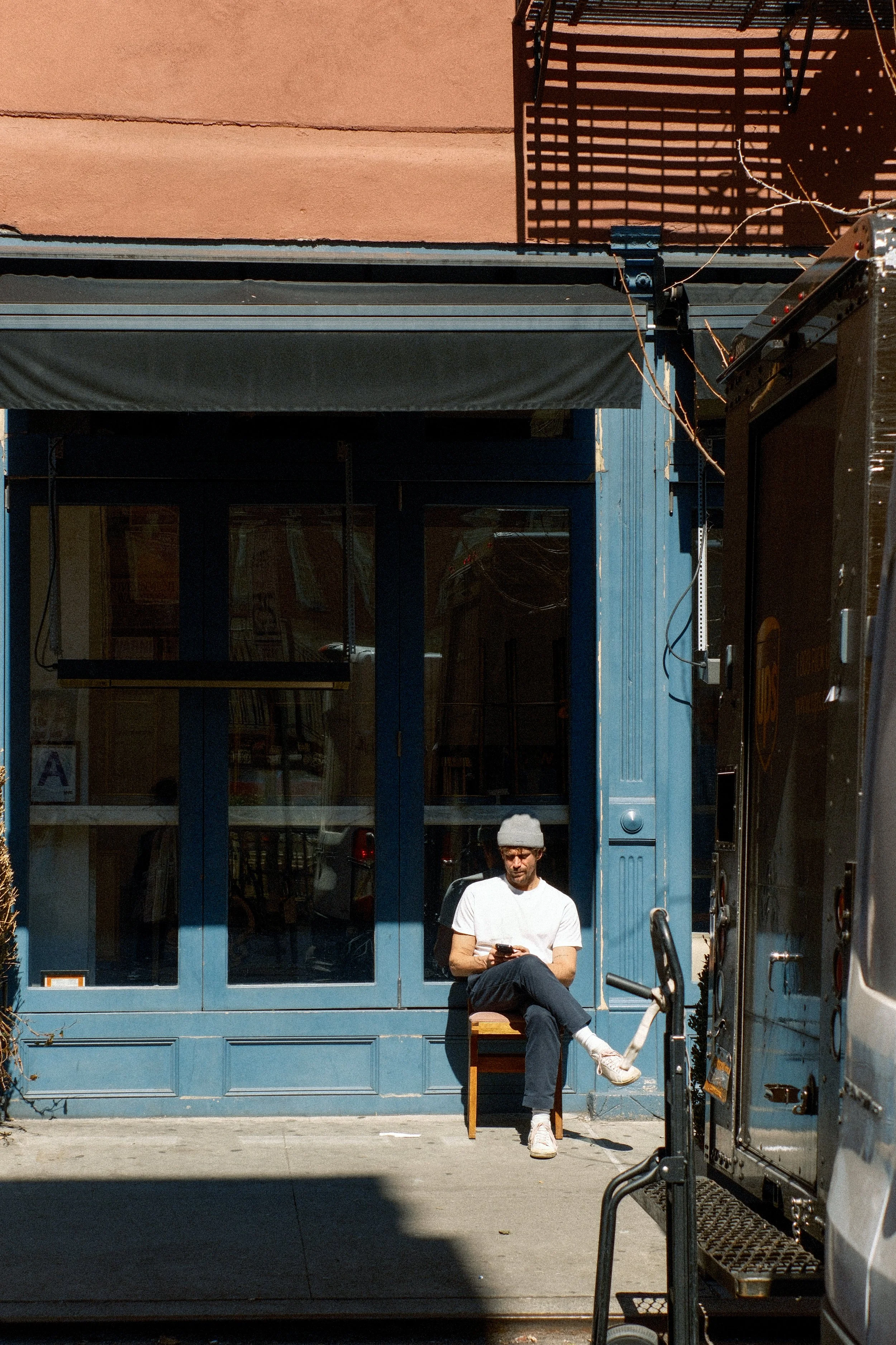A man wearing a gray beanie, white t-shirt, black pants, and white sneakers sitting on a chair outside a blue storefront, looking at his phone.