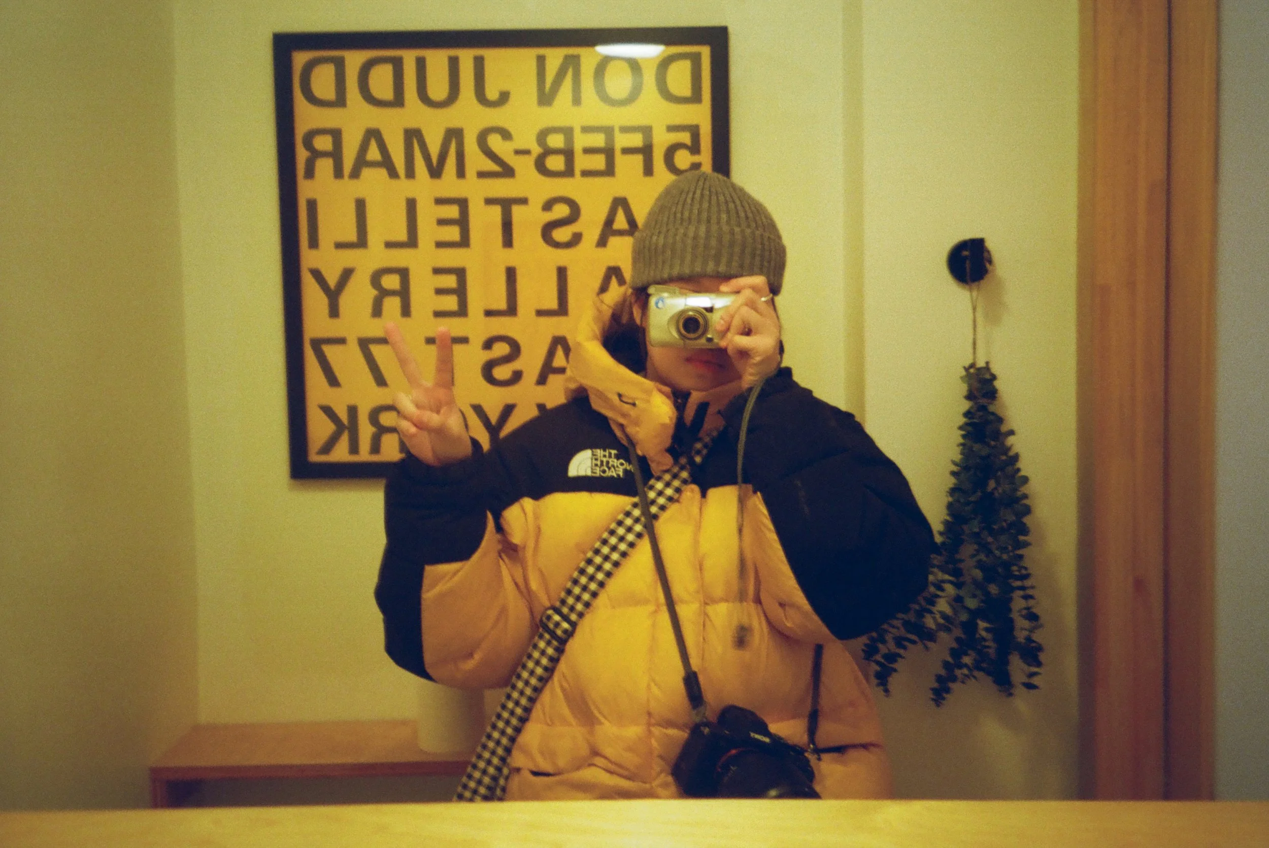 A person taking a mirror selfie, wearing a brown beanie, a black and yellow jacket, making a peace sign with their right hand, and holding a camera with their left hand. Behind them is a yellow wall with a framed eye chart and a small decorated Christmas tree.