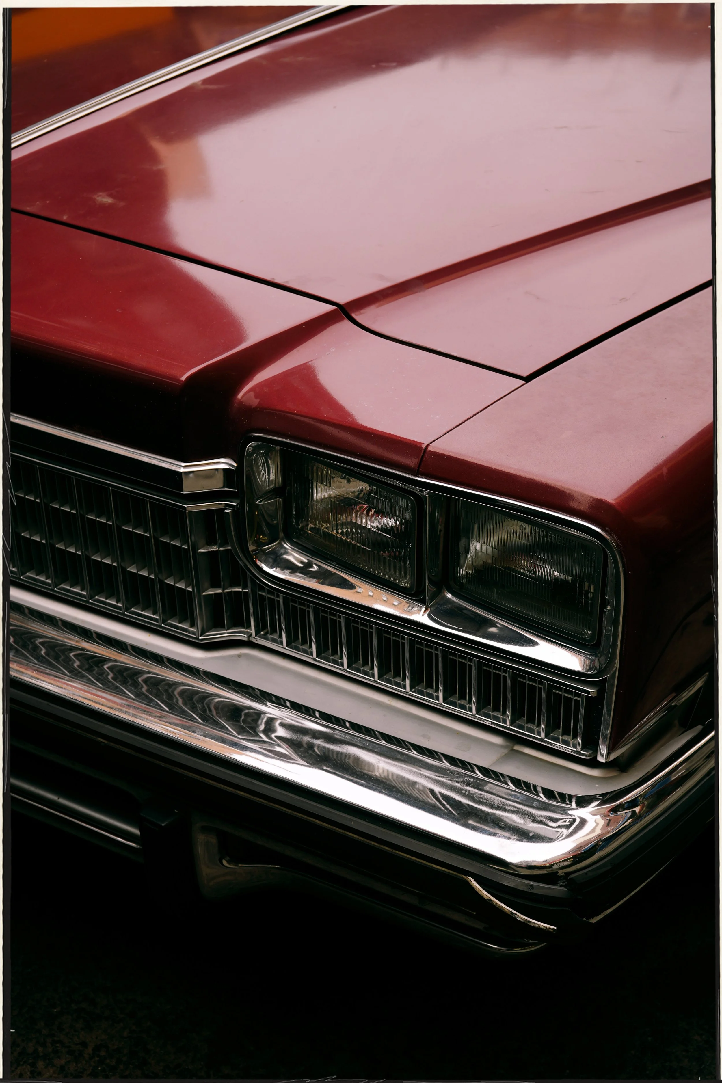 Close-up of the front of a vintage burgundy Cadillac car, showing the hood, grille, and headlight, with reflections on the chrome bumper.