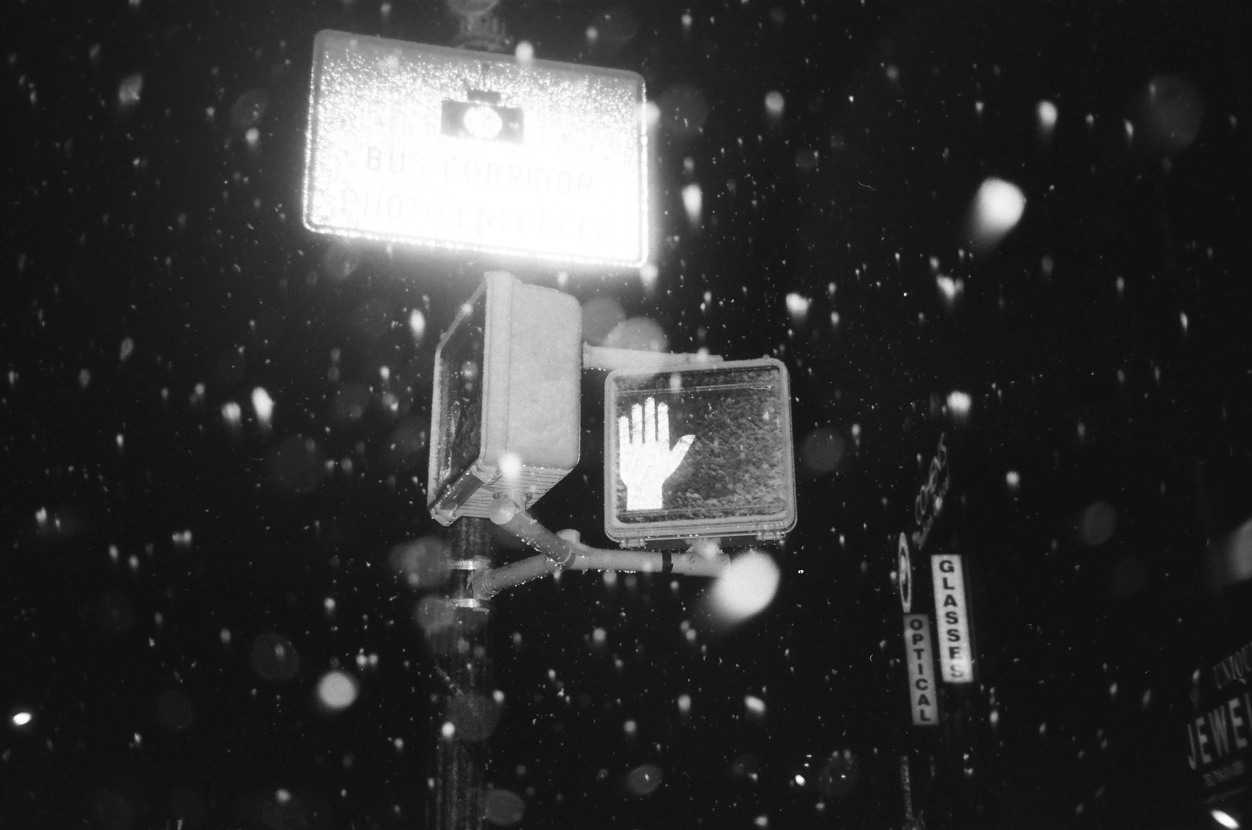 A parking lot during a snowstorm with a weather station and a traffic signal visible in the foreground.