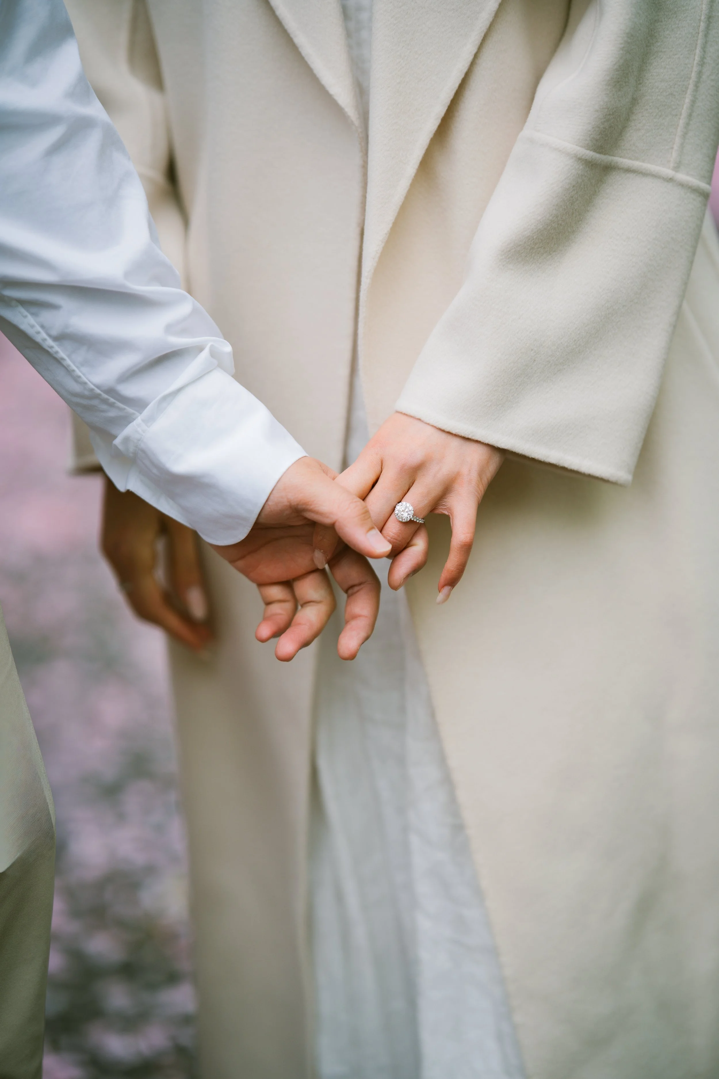 Two people holding hands, one wears a white shirt with a ring, the other wears a beige coat.