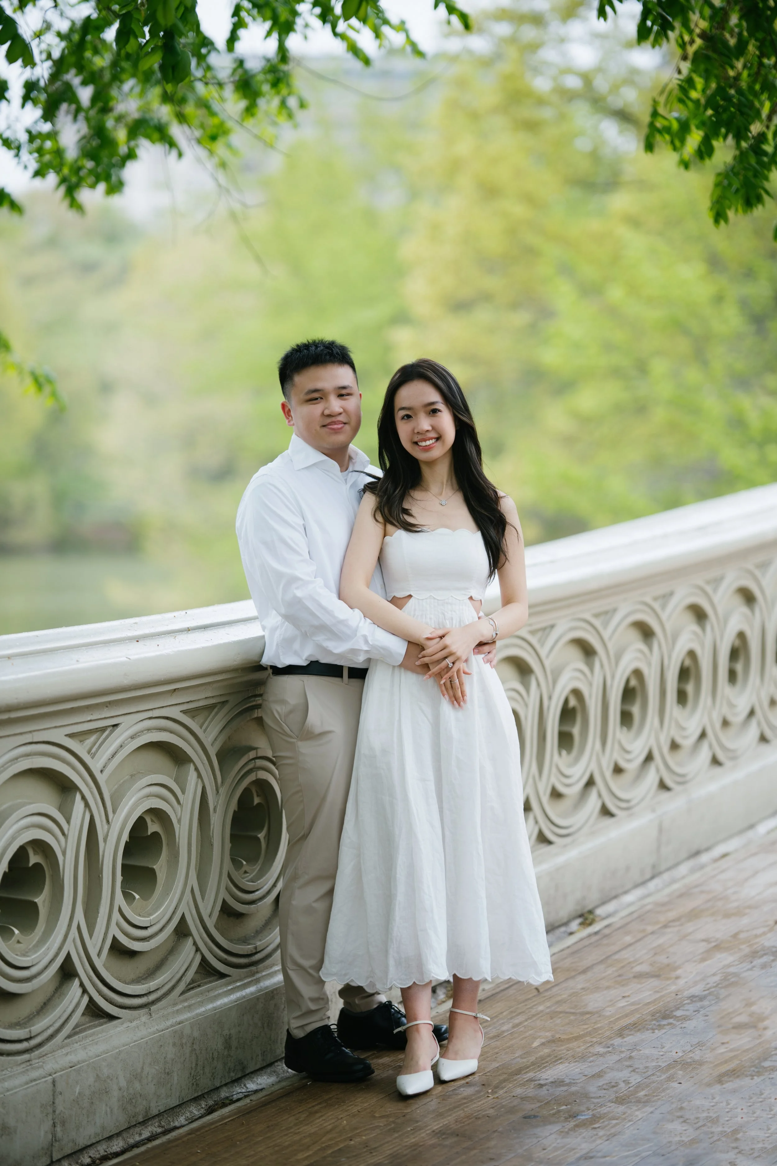 A young couple stands on a bridge with ornate railing, outdoors in a park-like setting, dressed in casual light-colored clothing, smiling at the camera, surrounded by green trees.