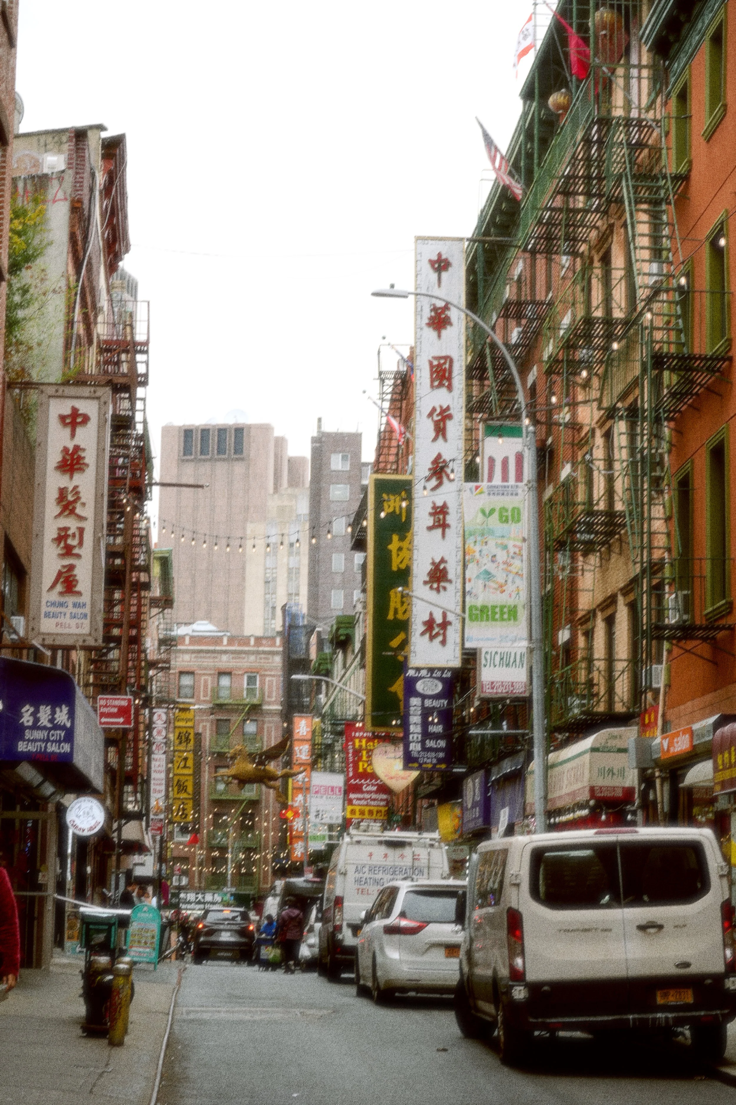 A busy city street area with cars parked on both sides, multicolored buildings with Chinese signs, and a mix of pedestrians and storefronts. Fire escape ladders are attached to the buildings, and the street is lined with shops and small businesses.