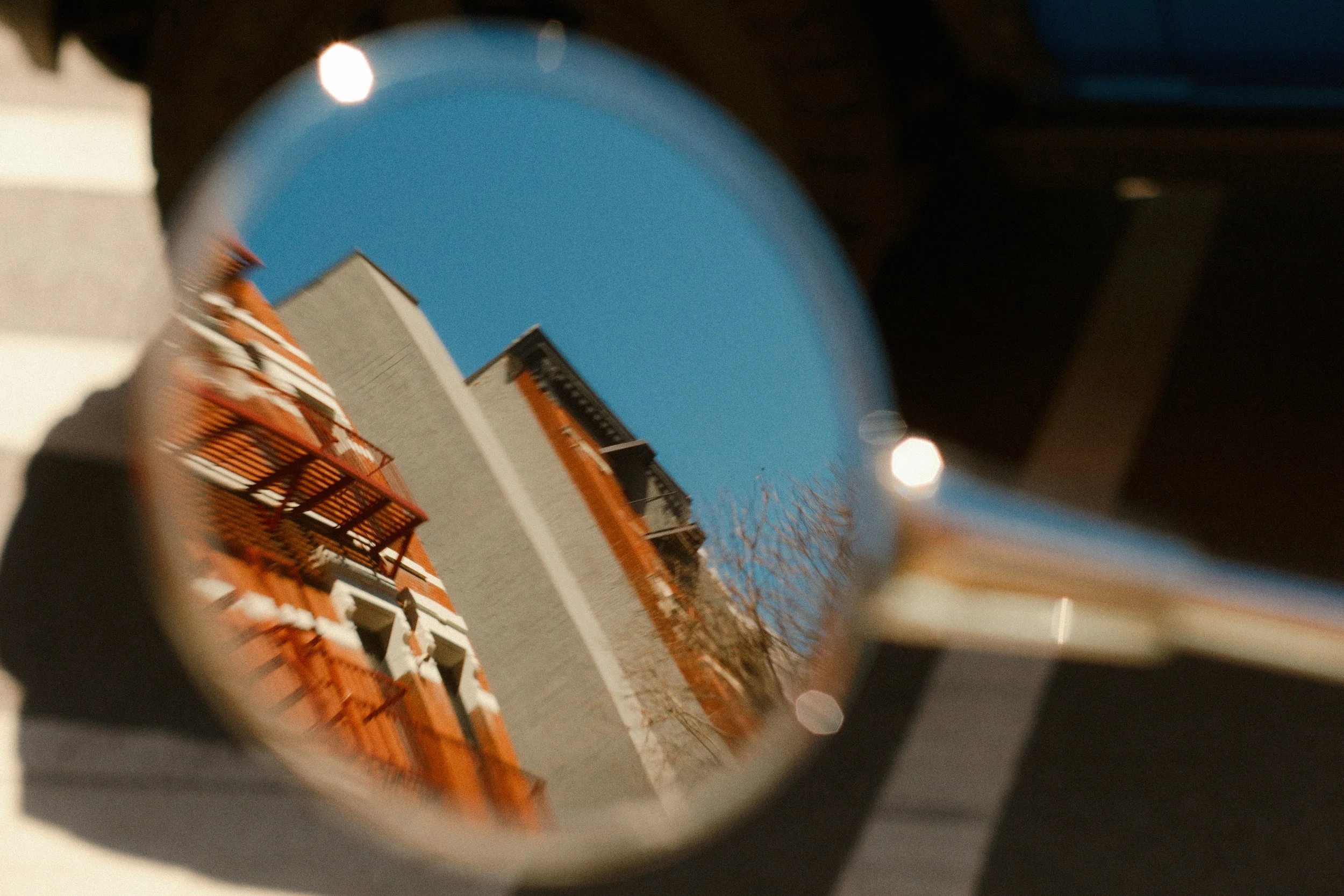 View of a building with orange accents seen through a circular object, possibly a telescope or a camera lens, with bokeh lights and a clear blue sky in the background.