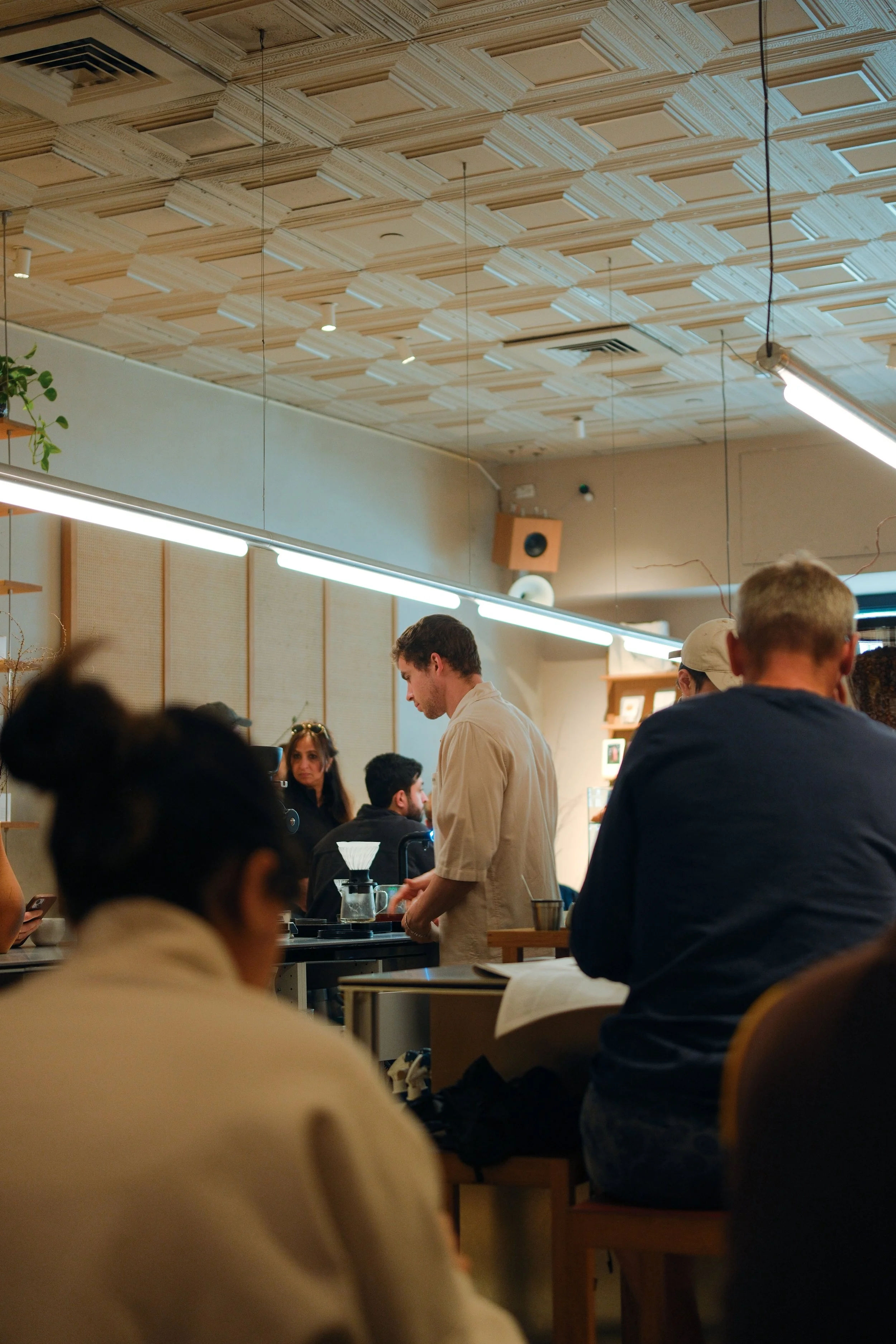 A group of people inside a cafe or restaurant, with a focus on a barista or worker in a beige shirt preparing a drink behind the counter, surrounded by customers. The ceiling has a decorative pattern with hanging lights, and there's a potted plant on