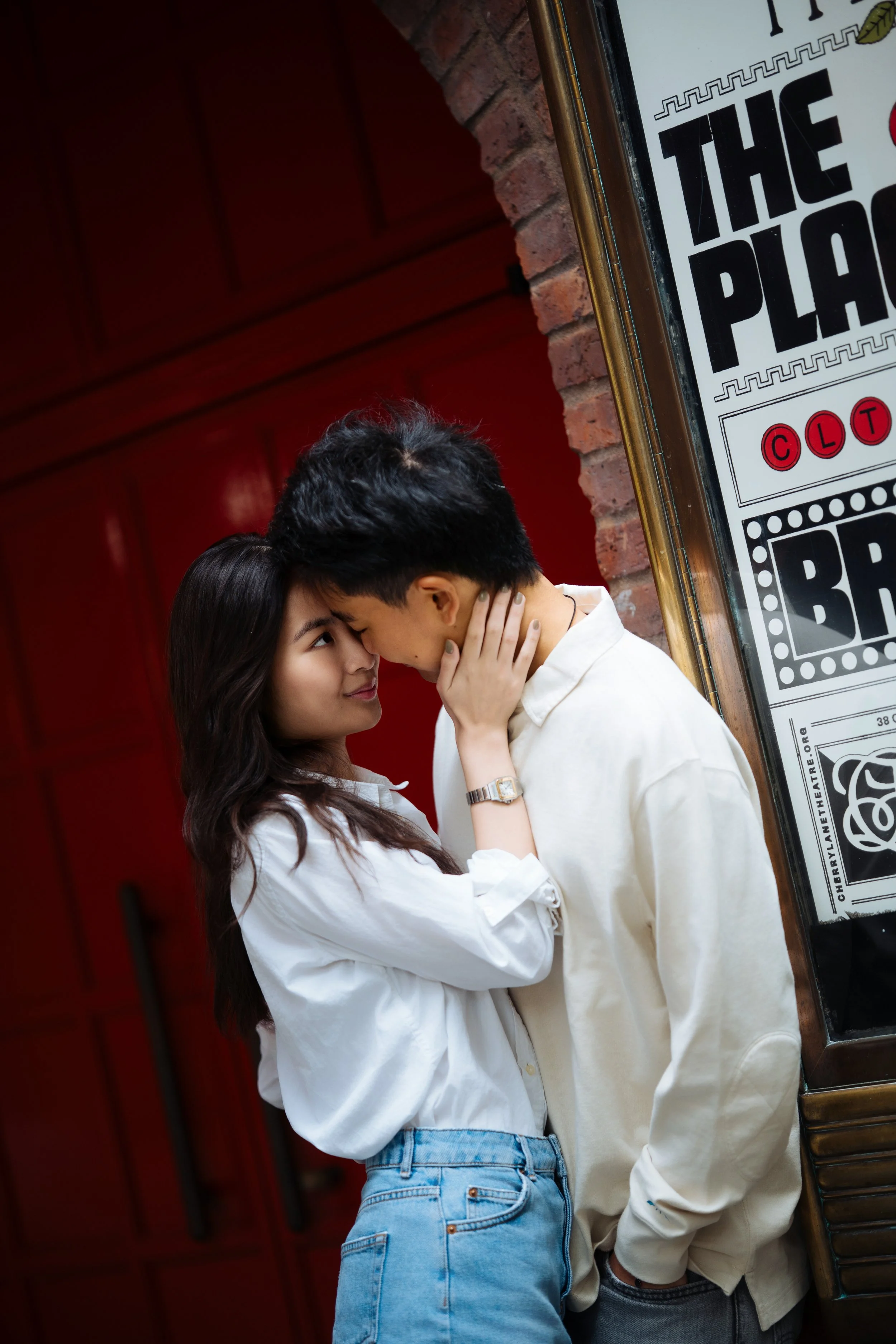 A couple standing close, touching foreheads and gazing into each other's eyes near a brick wall and a framed poster.