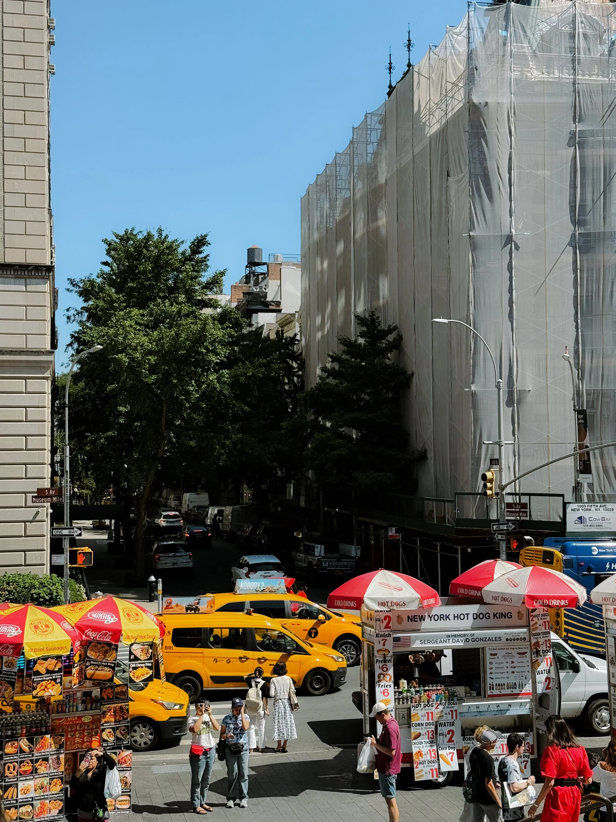 City street scene with pedestrians, yellow taxis, and food vendors. A building under renovation is wrapped in scaffolding and tarp. People are walking and waiting at a crosswalk, with umbrellas and signage for hot dogs and other fast food.