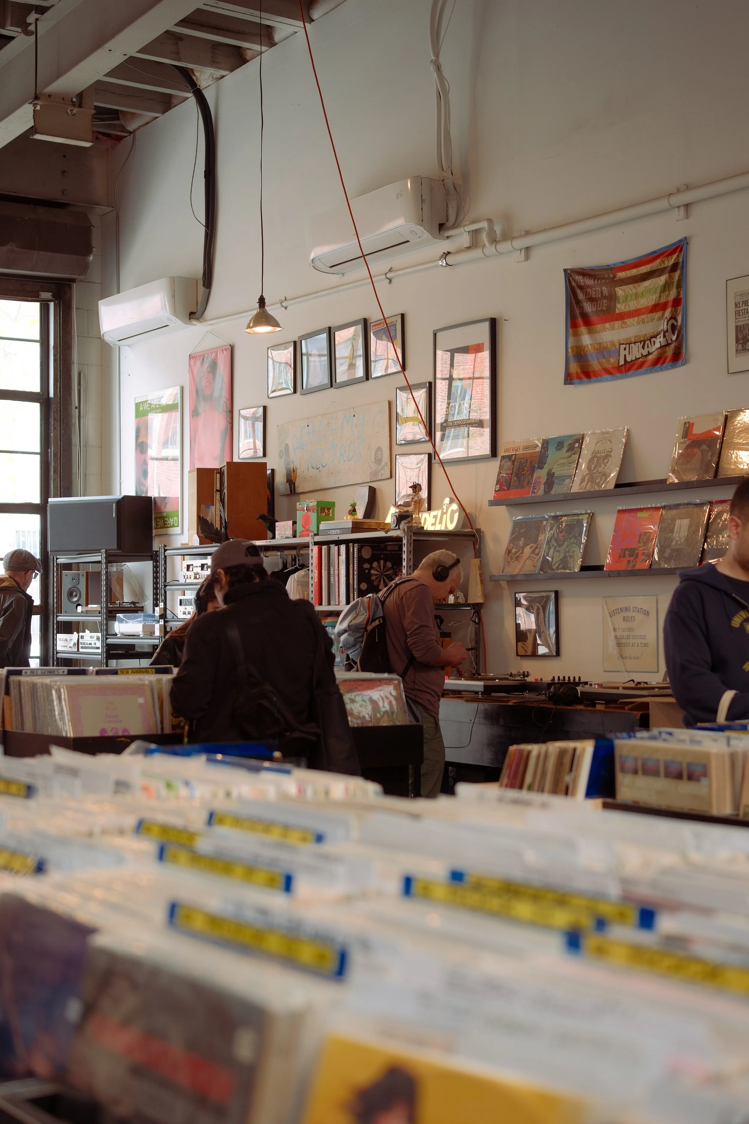 Inside a record store with shelves of vinyl records, framed artwork and posters on the white walls, and several people browsing. Large windows let in natural light.