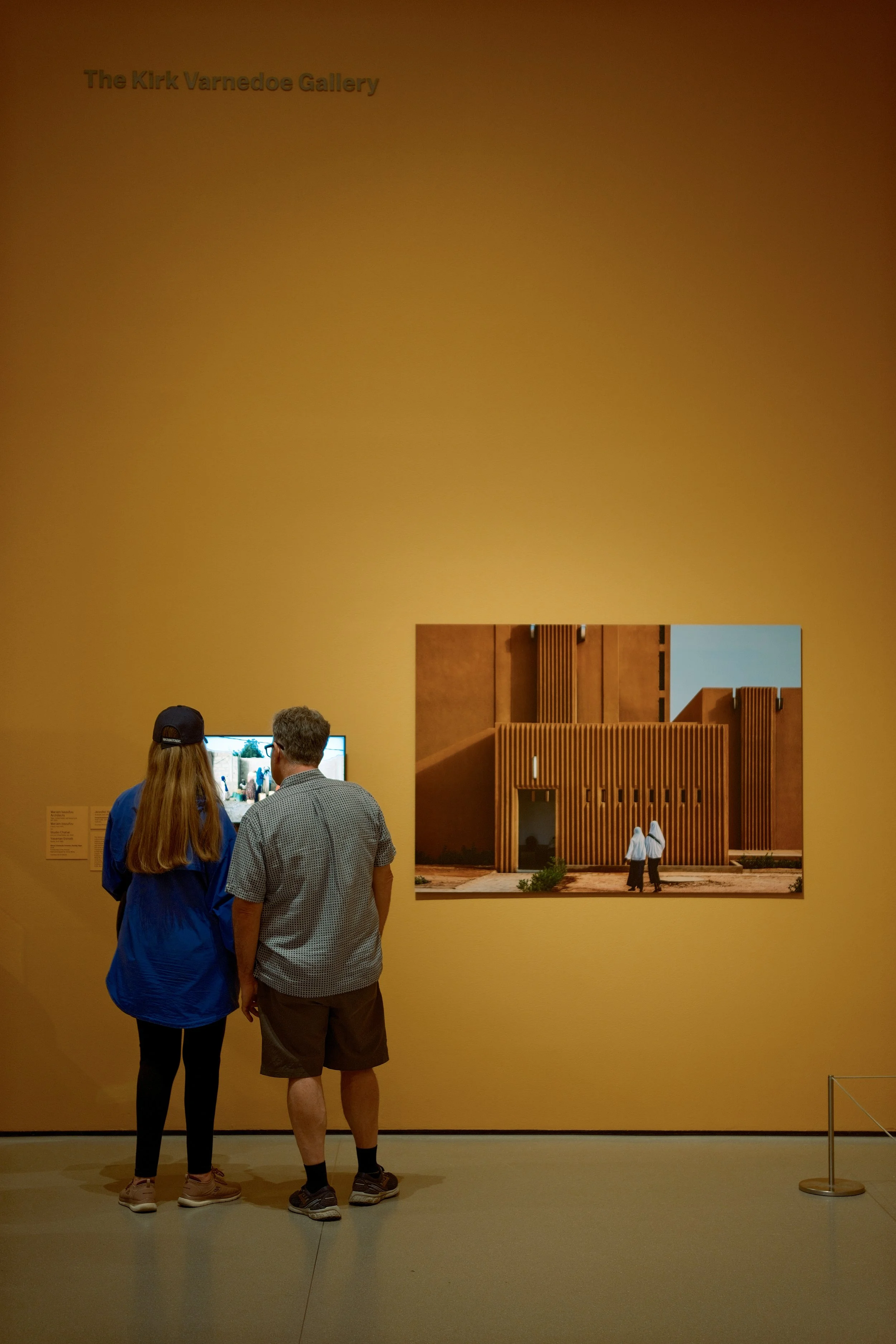 Two people, a woman with long hair and a man, standing in front of an art exhibit titled 'The Kirk Varnedoe Gallery' at the Kirk Varnedoe Gallery. They are looking at a screen with a digital display and an image of an architectural model on the wall 