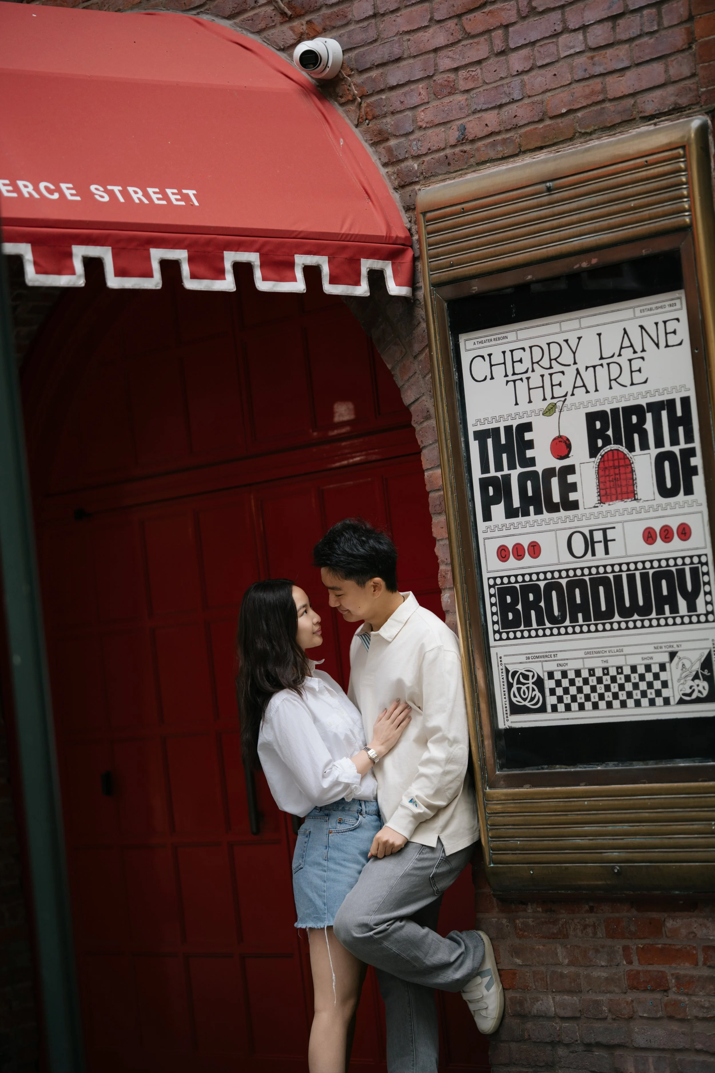 A young couple is standing close to each other outside a theater with a red brick wall background. The woman has long dark hair, is wearing a white shirt and denim shorts, and looks into the man's eyes. The man has short dark hair, is dressed in a white shirt and gray pants, leaning against the brick wall, and gazing back at the woman. They are positioned near a vintage theater marquee that advertises a show called "The Birth of Broadway."