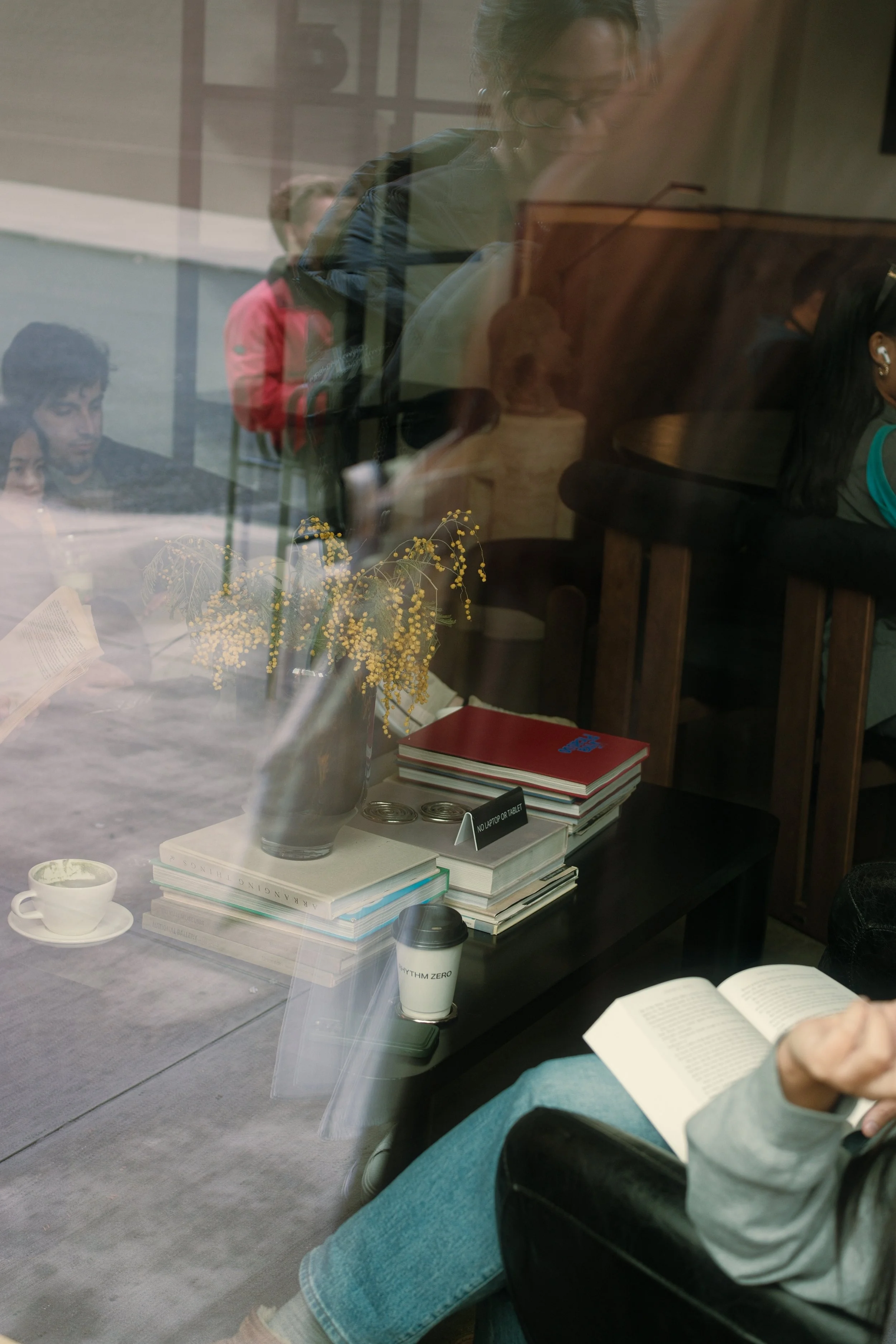 Photo of a person sitting at a table reading a book inside a cafe, viewed through a glass window with reflections of other people and objects outside.