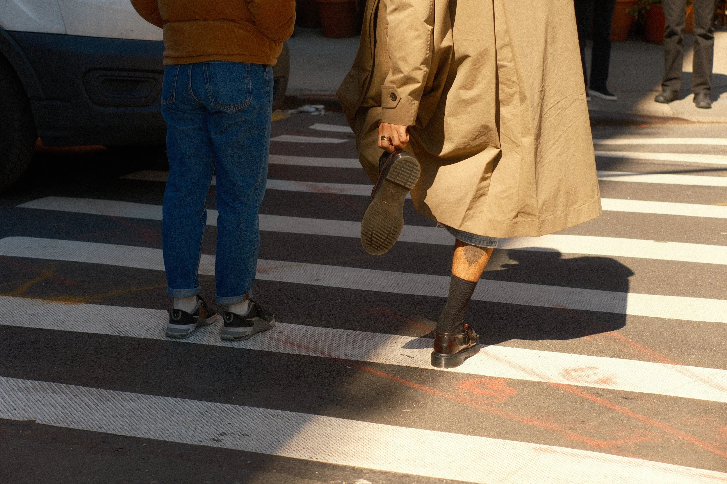 People crossing a pedestrian crosswalk, with focus on legs and feet, wearing casual clothing and shoes, with a parked vehicle and other pedestrians in the background.
